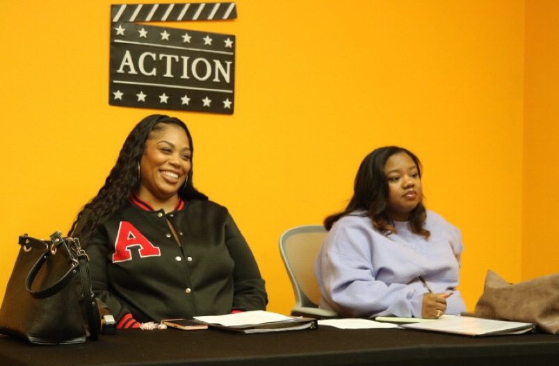Two women sit at a table with notebooks and papers, one smiling and the other looking serious, against a yellow wall.