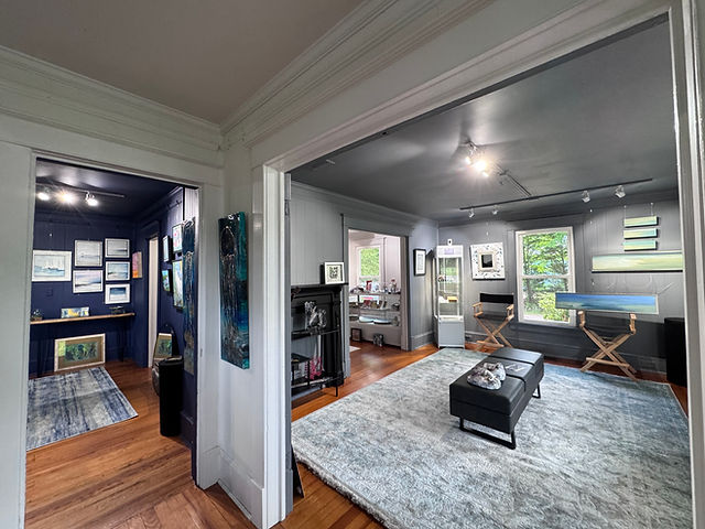 Living room with large windows, gray walls, a black ottoman, and a gray rug, connected to a smaller room with a blue wall.