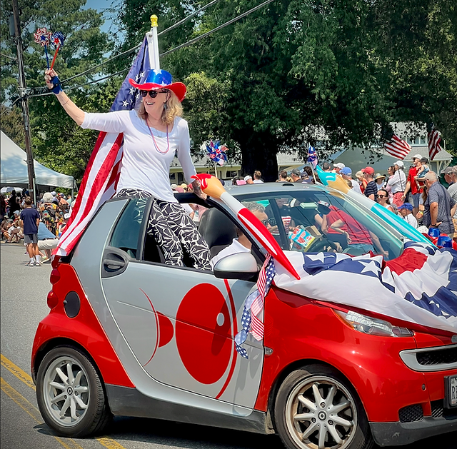 Person dressed as Uncle Sam standing through a decorated small car during parade, holding an American flag, with crowd and trees in background.