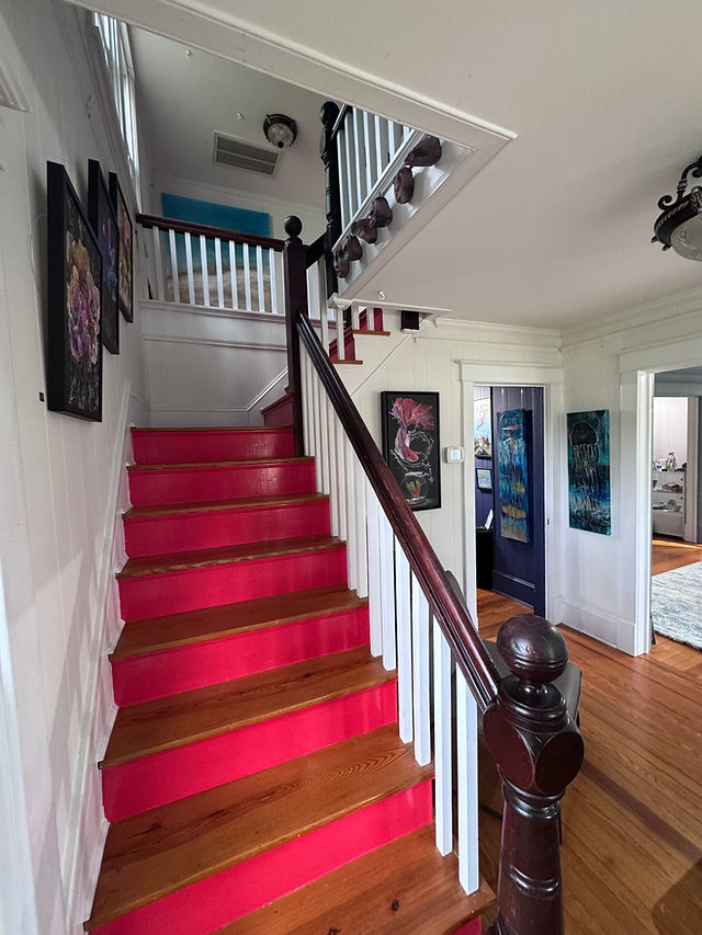 Interior view of a staircase with pink carpeted steps, wooden handrail, and framed artwork on walls, leading to upper floor.