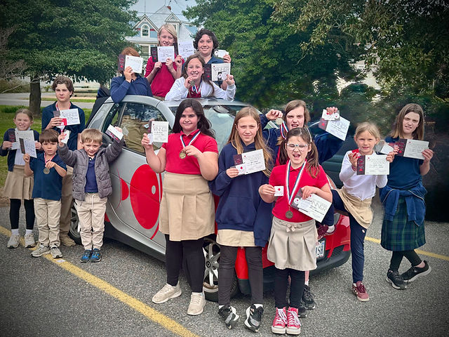 Group of children and adults holding certificates in front of a car with a red heart symbol, outdoors with trees and a building.