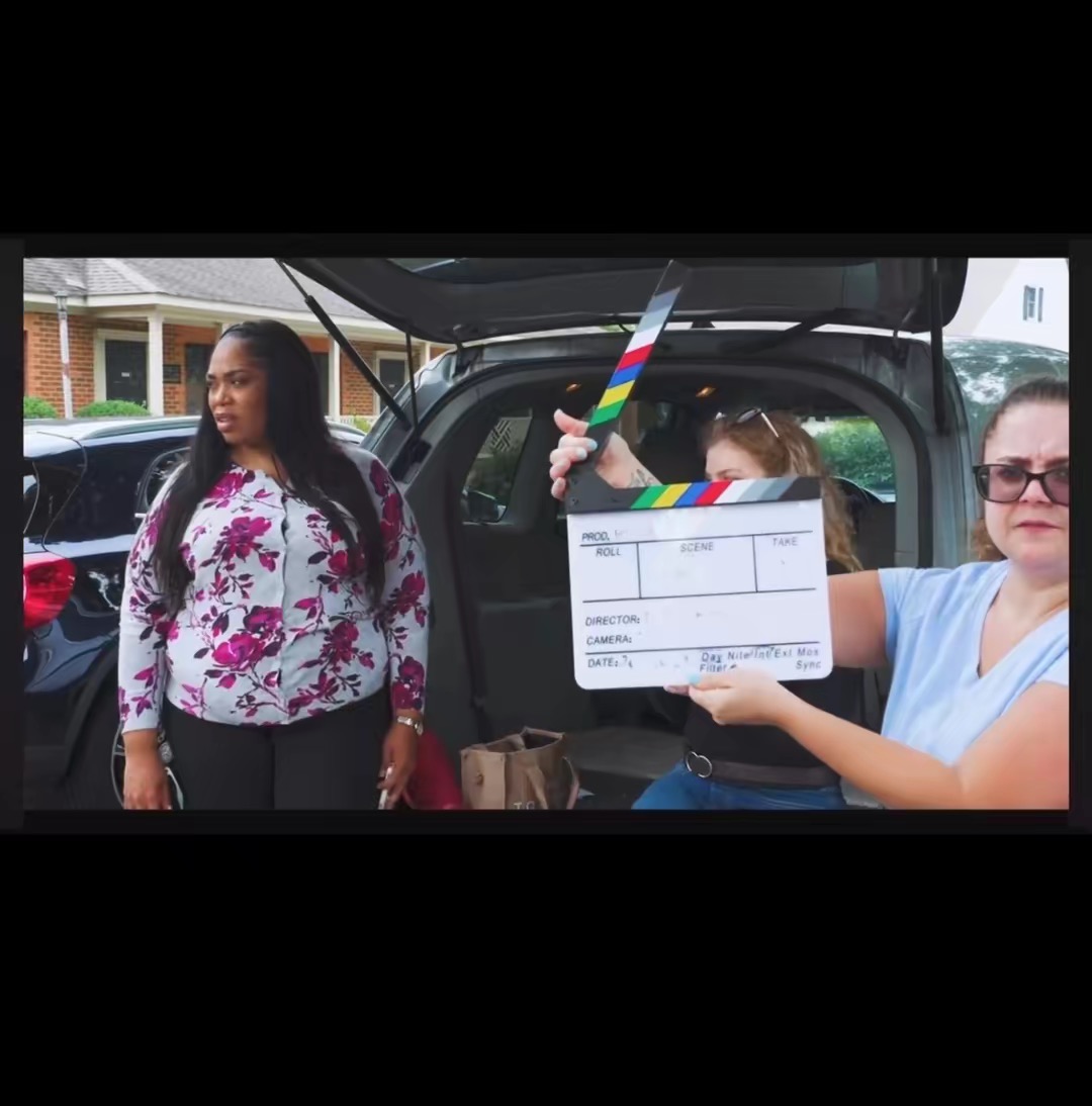 A woman in a floral blouse stands outside near a car, while another woman holds a clapperboard in front of her.