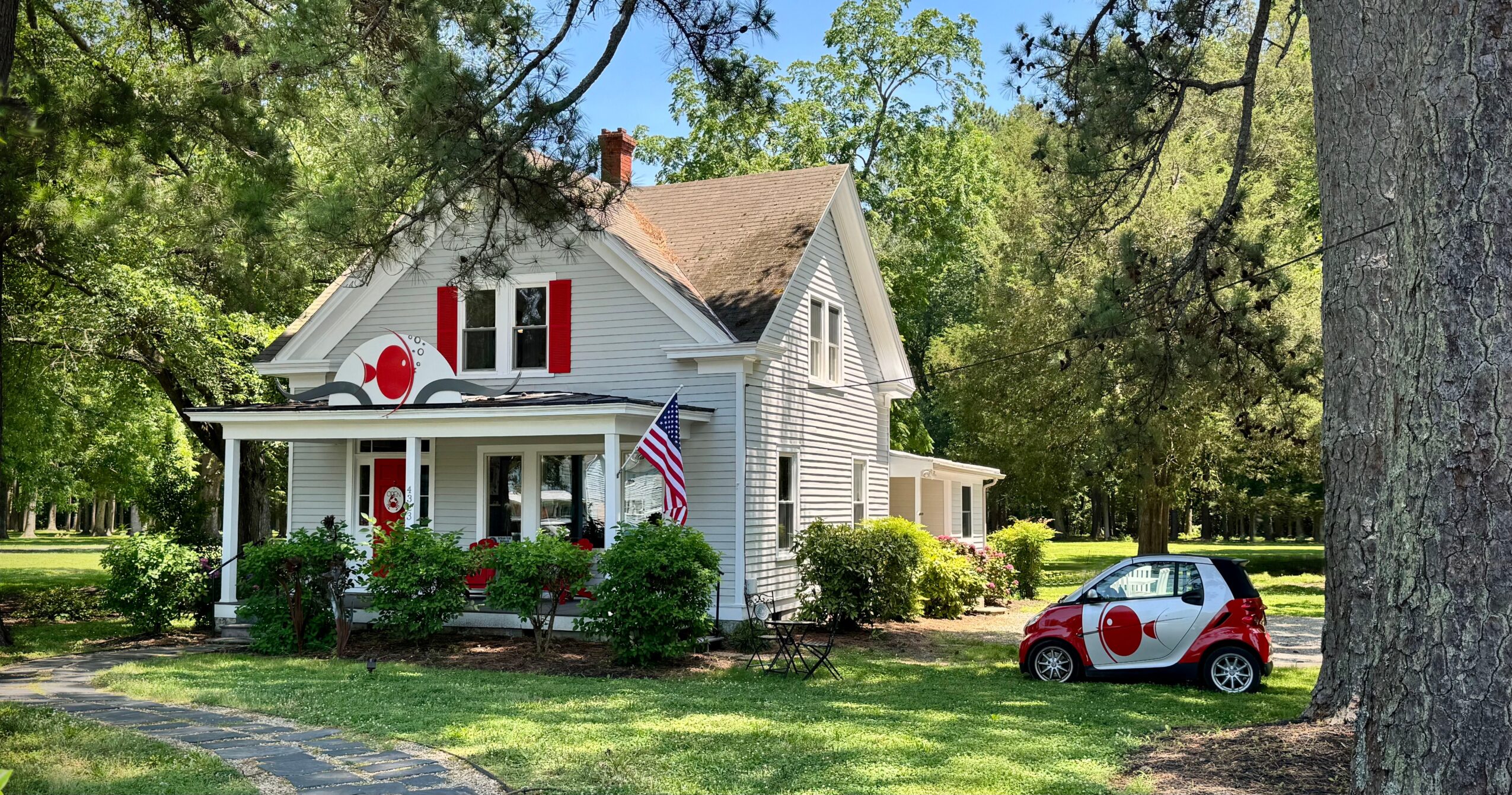 White house with red and white decorations, American flag, small car with red and white design, surrounded by trees and greenery.