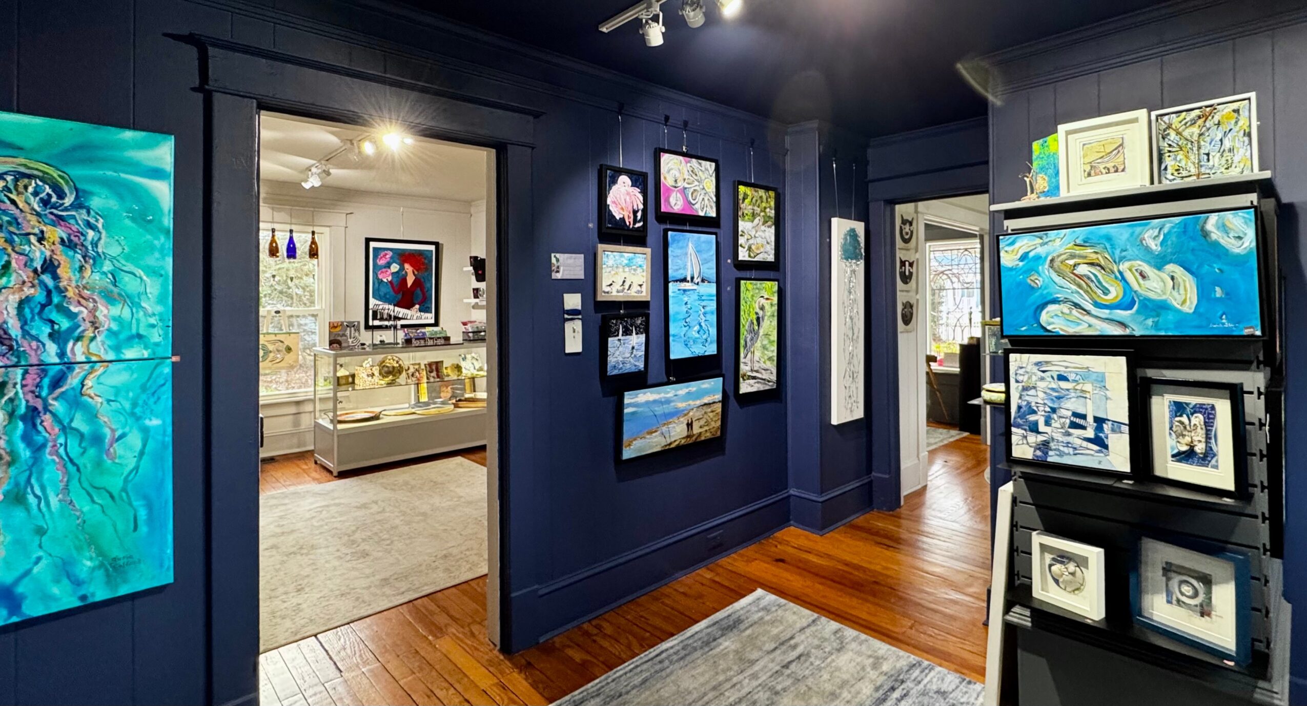 Art gallery with blue walls displaying paintings and a display case with artwork, hardwood floor, and a doorway to another room.