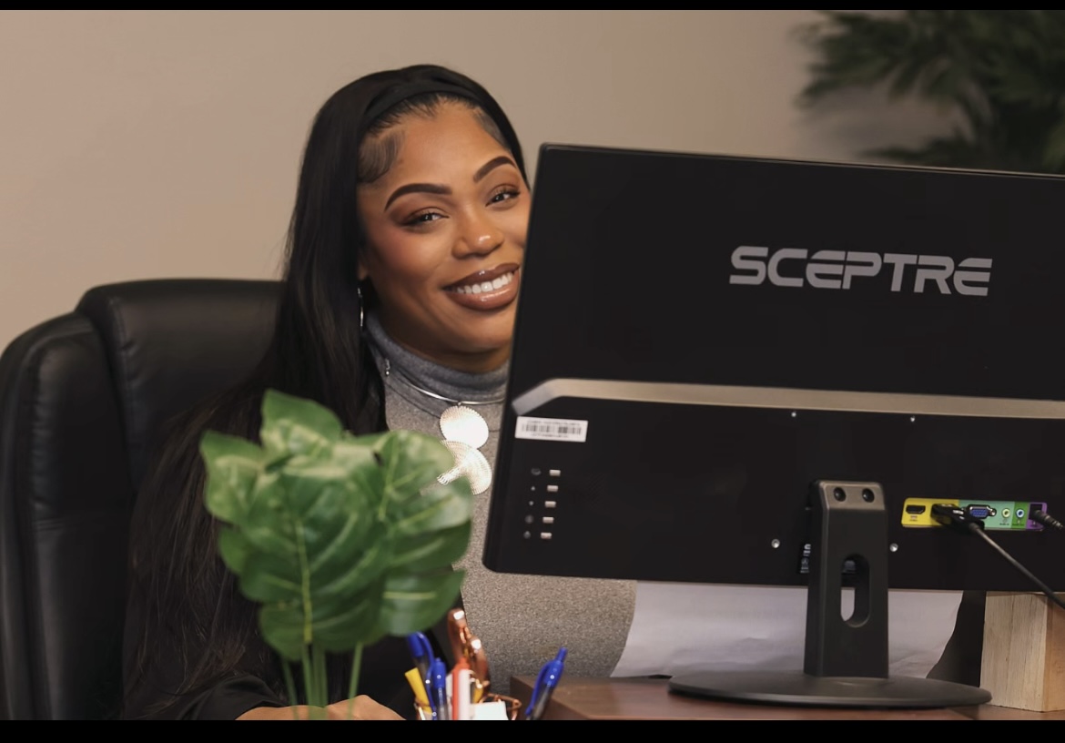 Woman smiling at her desk with a computer monitor, plant, and office supplies, in an indoor setting.