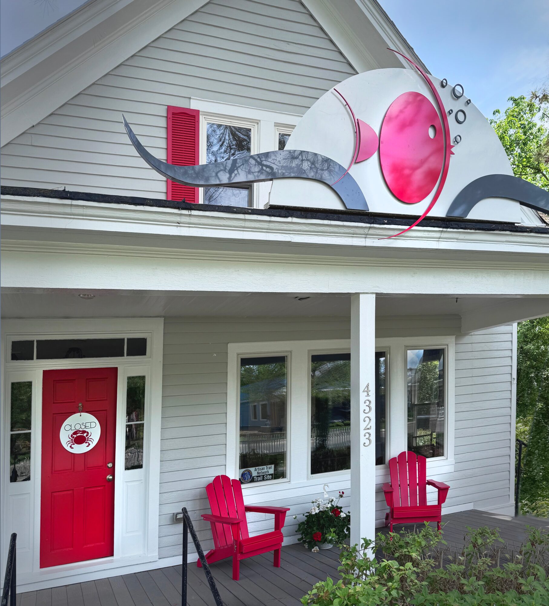 Large fish sculpture on house roof, with pink and white colors, and two red chairs in front of house, flowers, and house number 4023.
