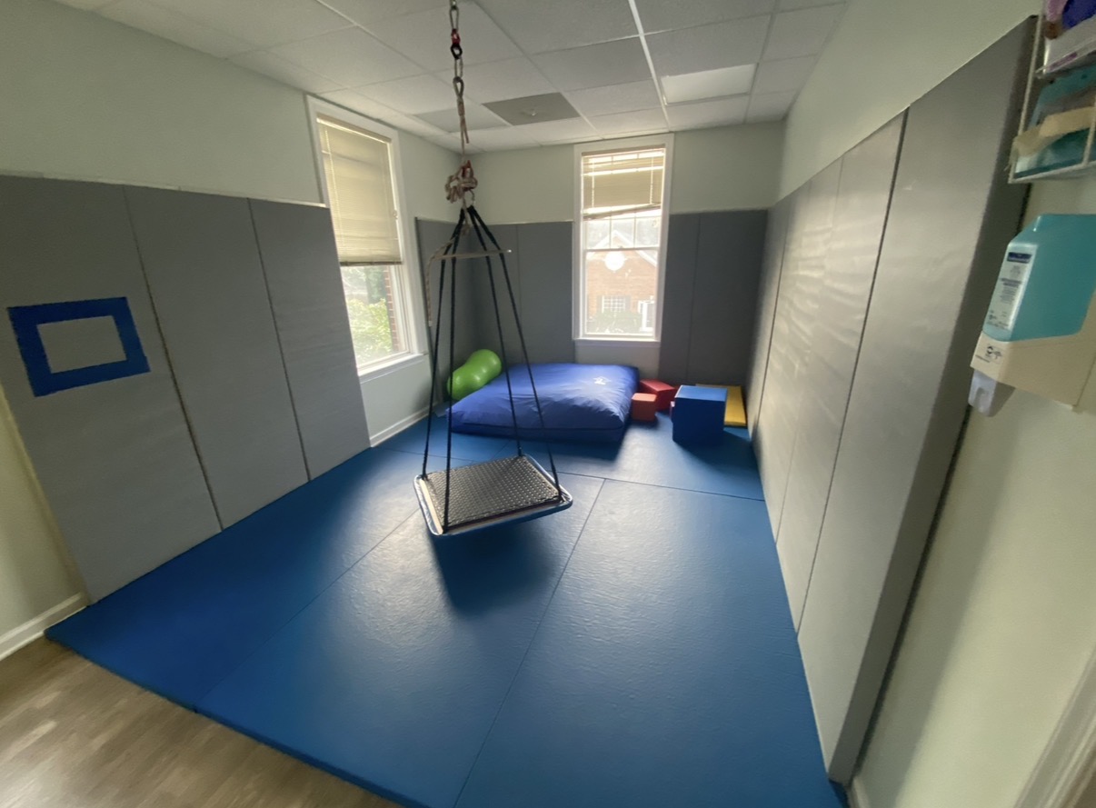 Indoor play area with a swing, padded mat, and soft toys, illuminated by natural light from windows.