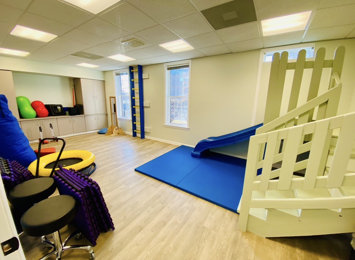 Indoor play area with a slide, soft mat, and various colorful equipment, bright room with windows and wooden flooring.