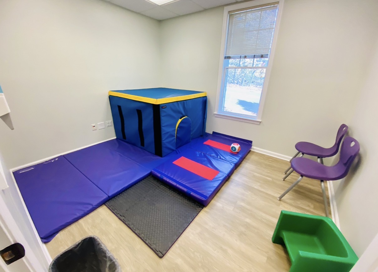Room with gym mats, a padded cube, a window, two purple chairs, and a green container, with light-colored walls and wooden floor.