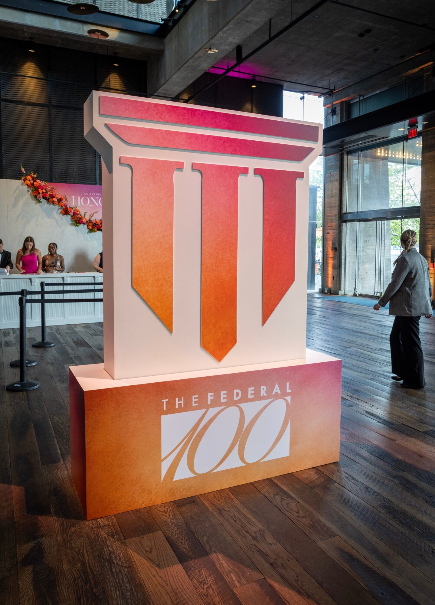 Large display with a stylized Roman numeral and text celebrating the federal 100th anniversary, inside a modern building with glass walls.