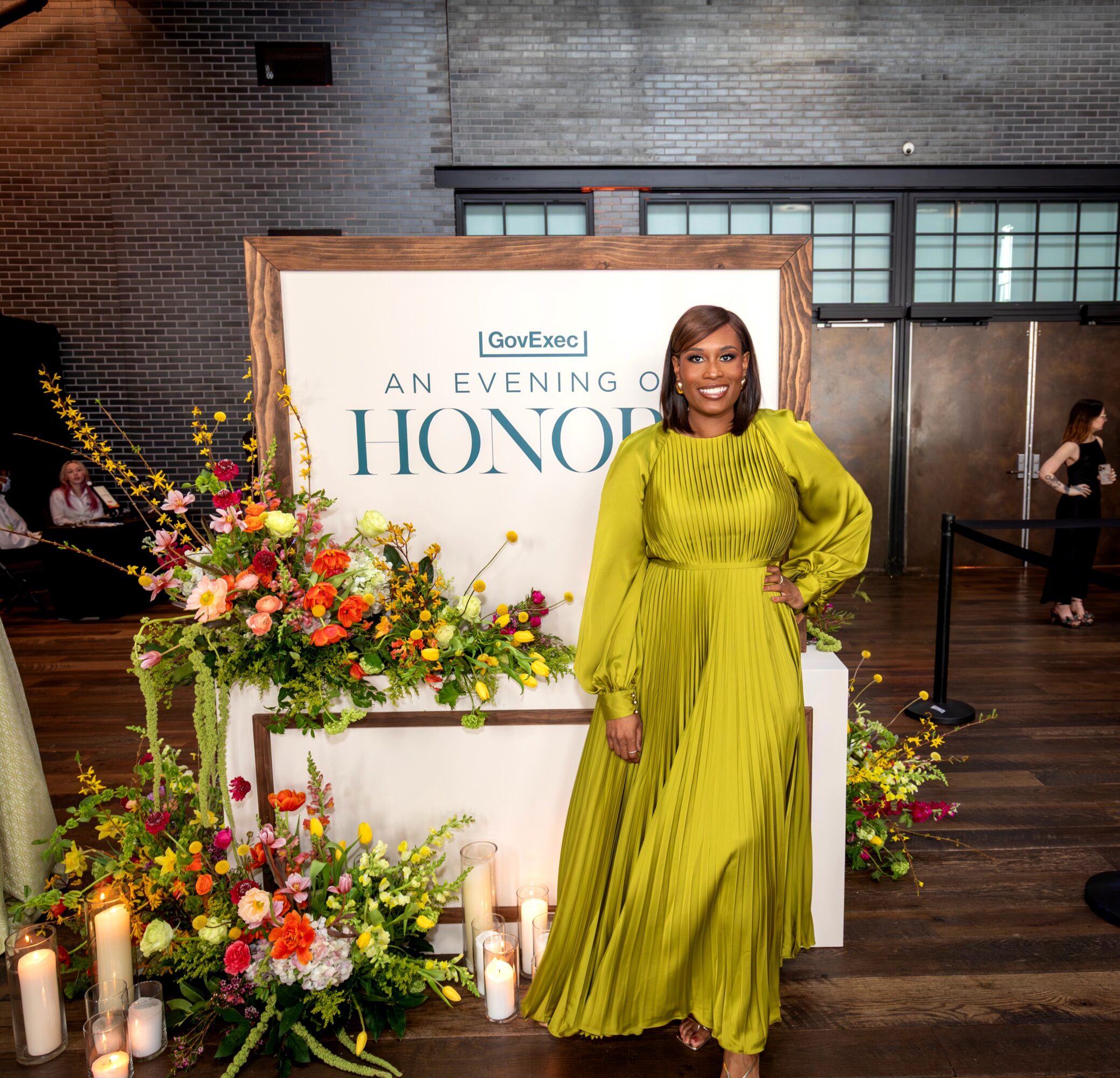 Woman in a bright yellow dress standing next to a floral arrangement and candles at an event, smiling.