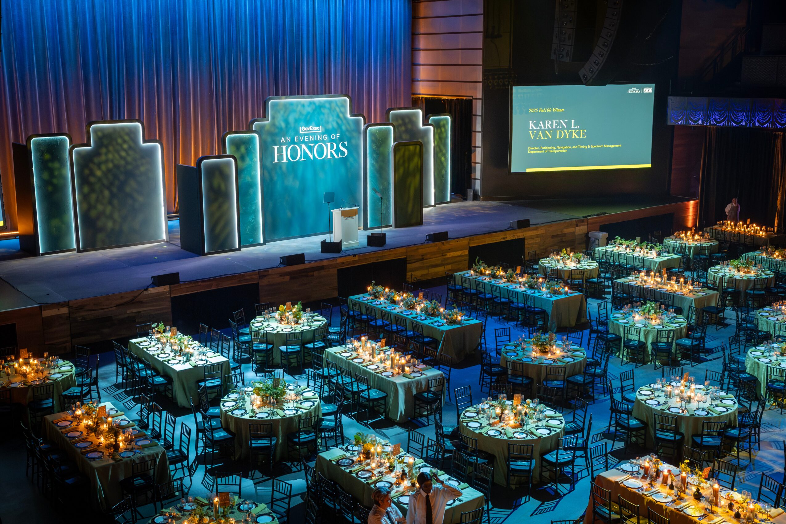 Banquet hall with round tables set for an event, decorated with candles and flowers, stage with banners and a screen in the background.