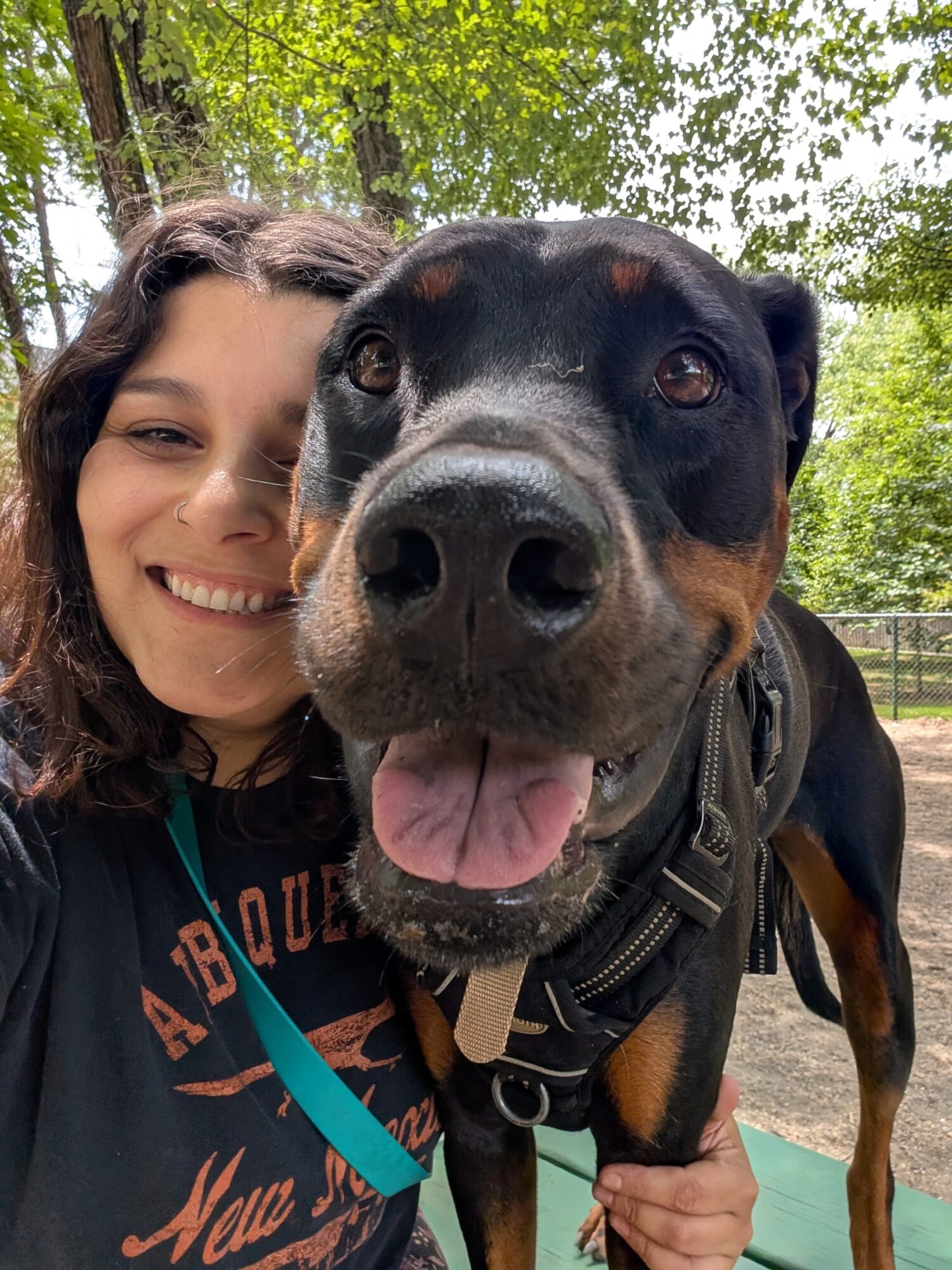 Person smiling outdoors with a large black and brown dog, both looking at the camera, trees in background.