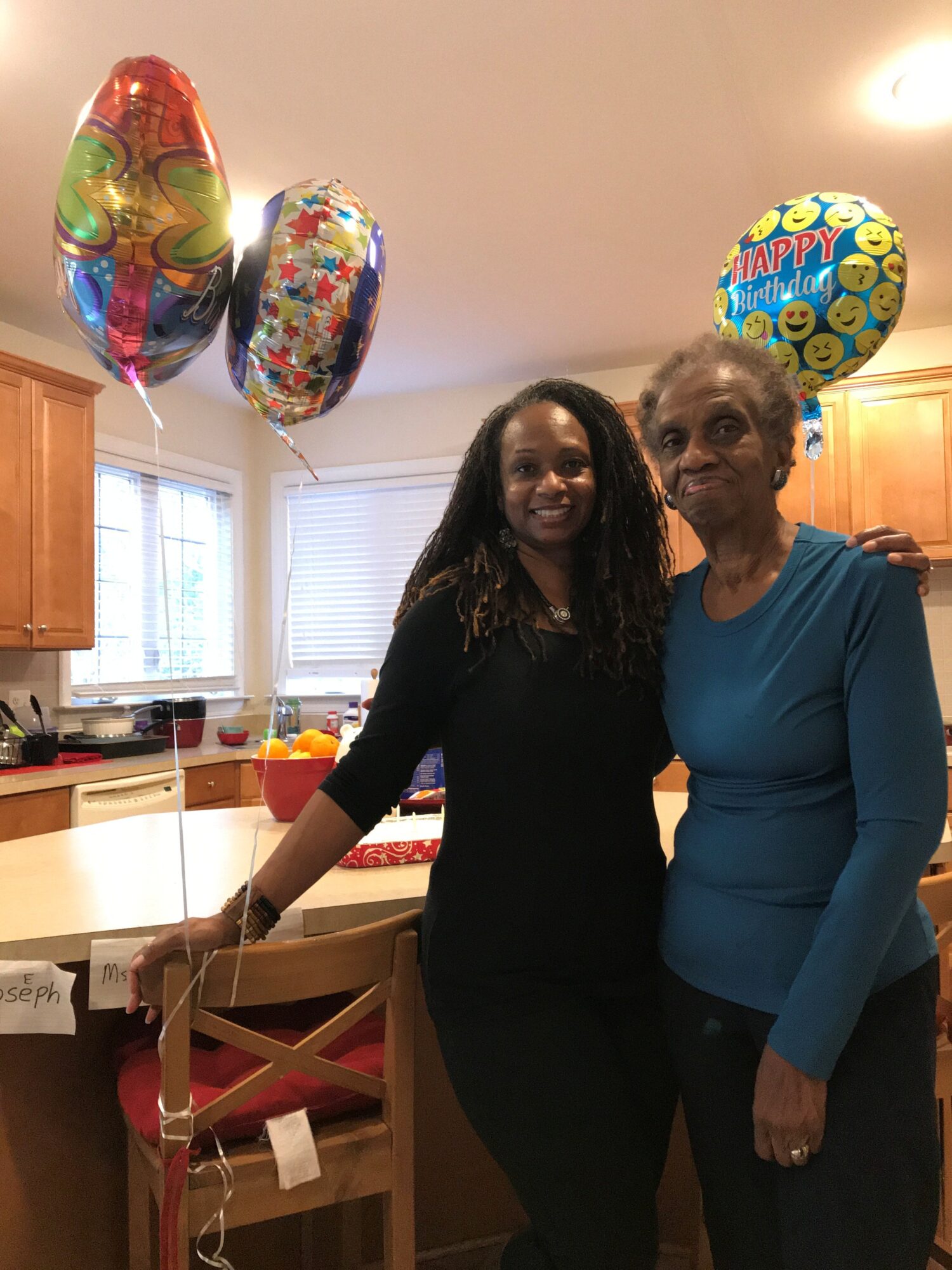 Two women standing in a kitchen with balloons, smiling, one younger with long dark hair, one older with short gray hair, both wearing long-sleeved tops.