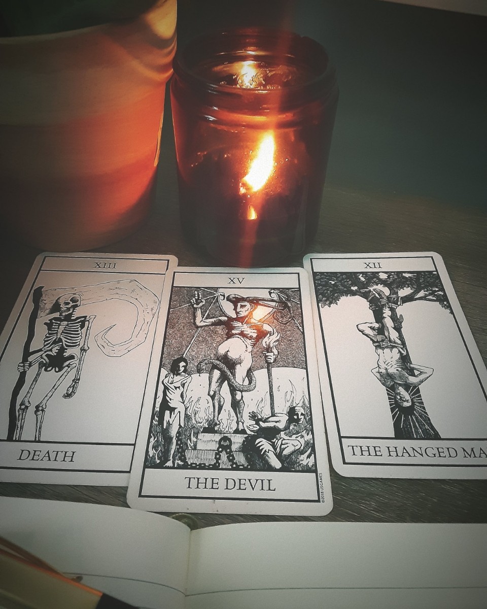 Three tarot cards labeled Death, The Devil, and The Hanged Man are laid out on an open book, with a lit candle nearby.