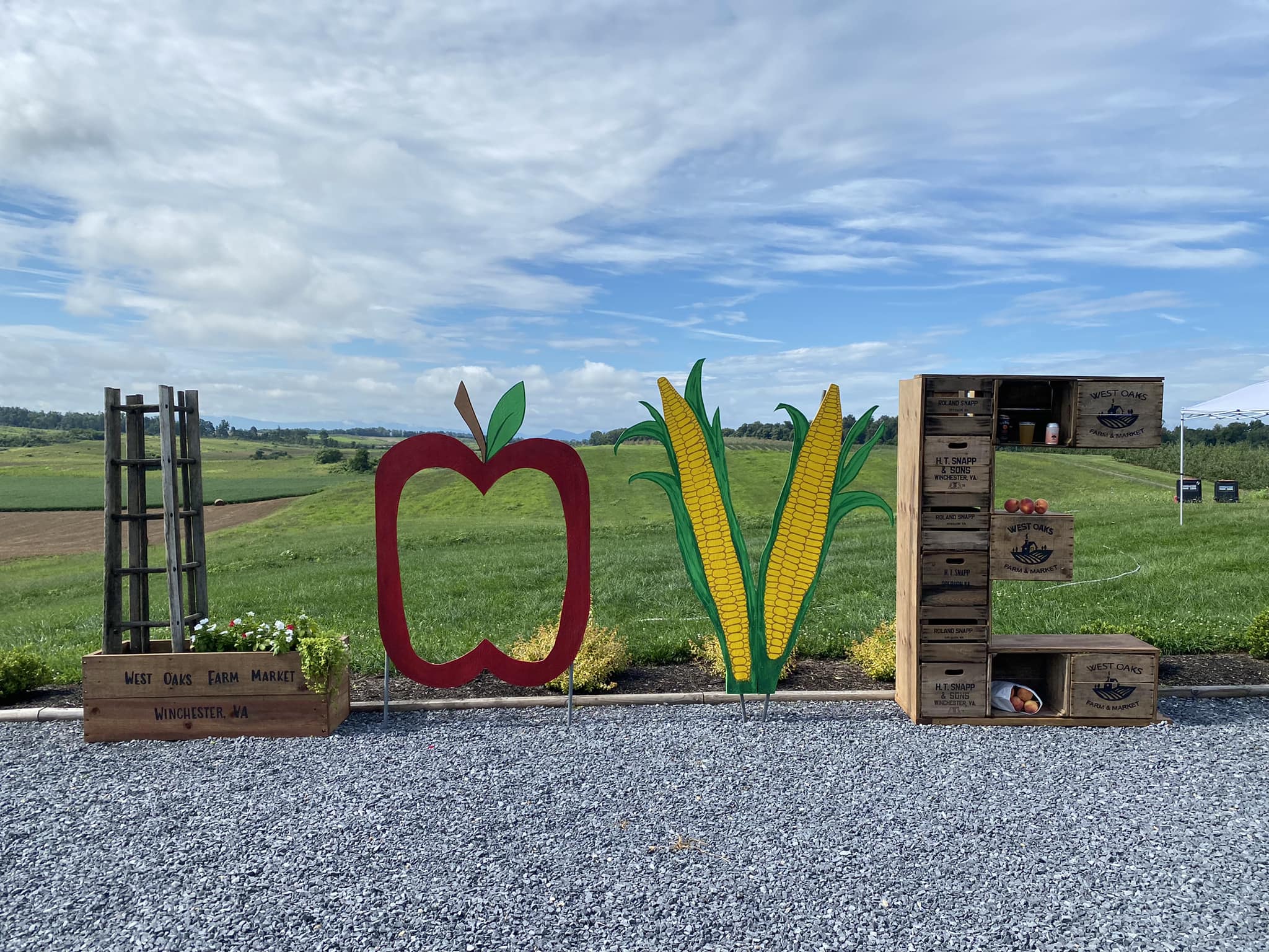 Outdoor display spelling 'CORN' with a red apple, corn cobs, and wooden crates on gravel against a field and sky