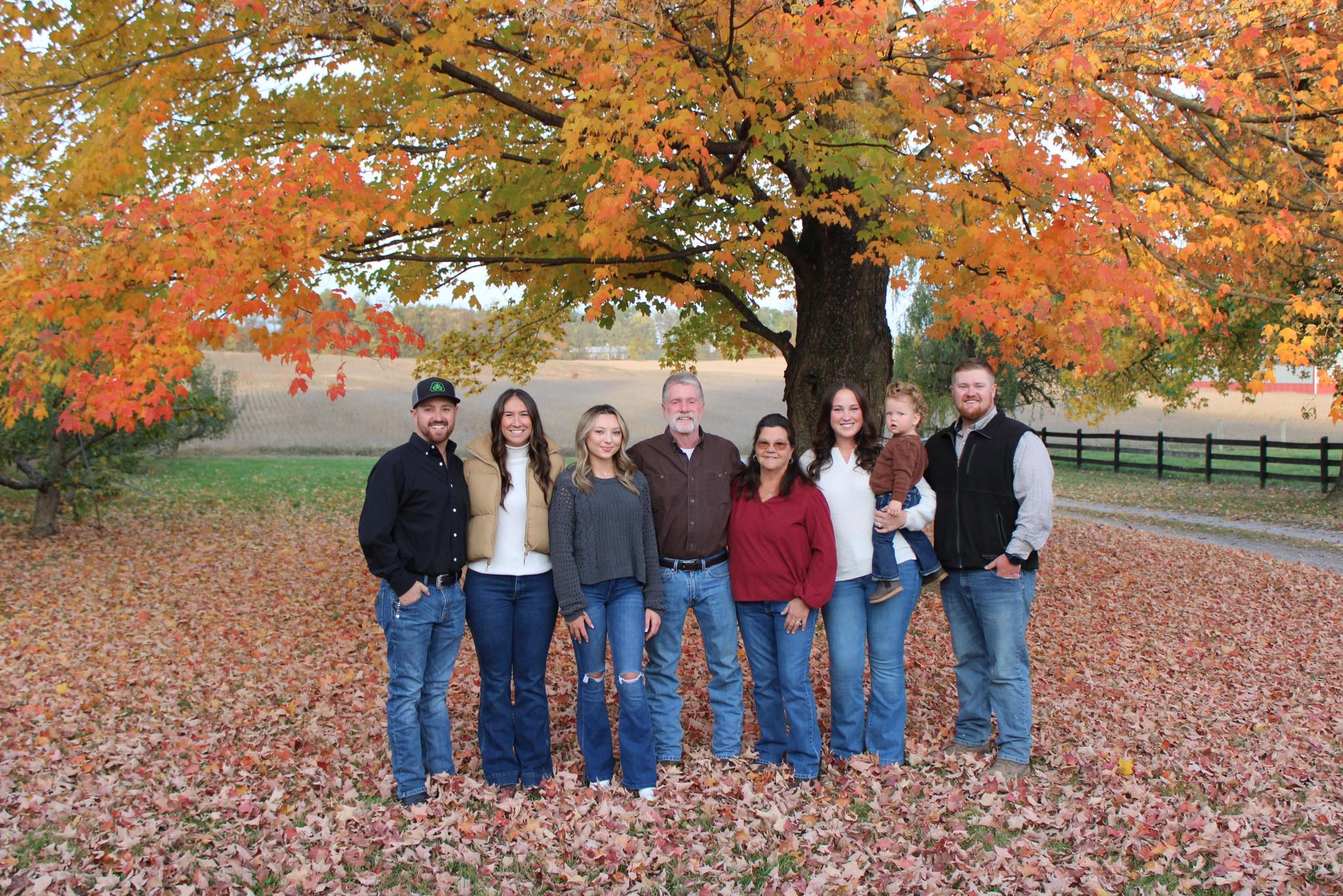 Group of seven people standing outdoors under a large tree with orange and yellow leaves, autumn scenery, and fallen leaves on the ground.