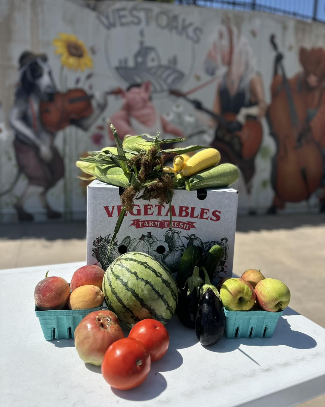 Display of fresh vegetables and fruits including watermelon, apples, eggplant, and zucchini in front of a mural with musicians.