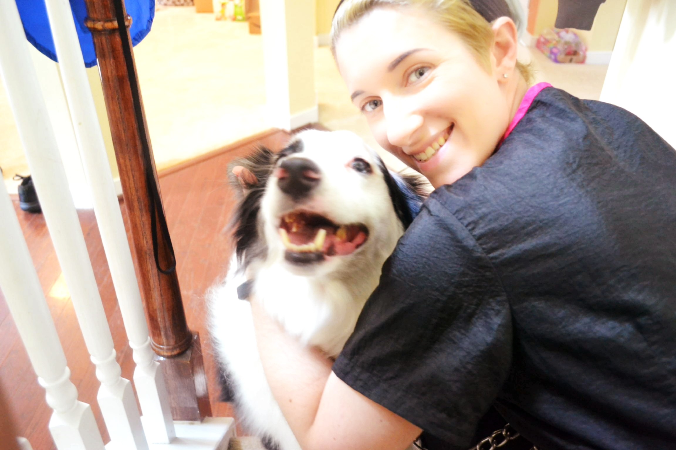 Young woman smiling and hugging a black and white dog indoors, with a staircase in the background.