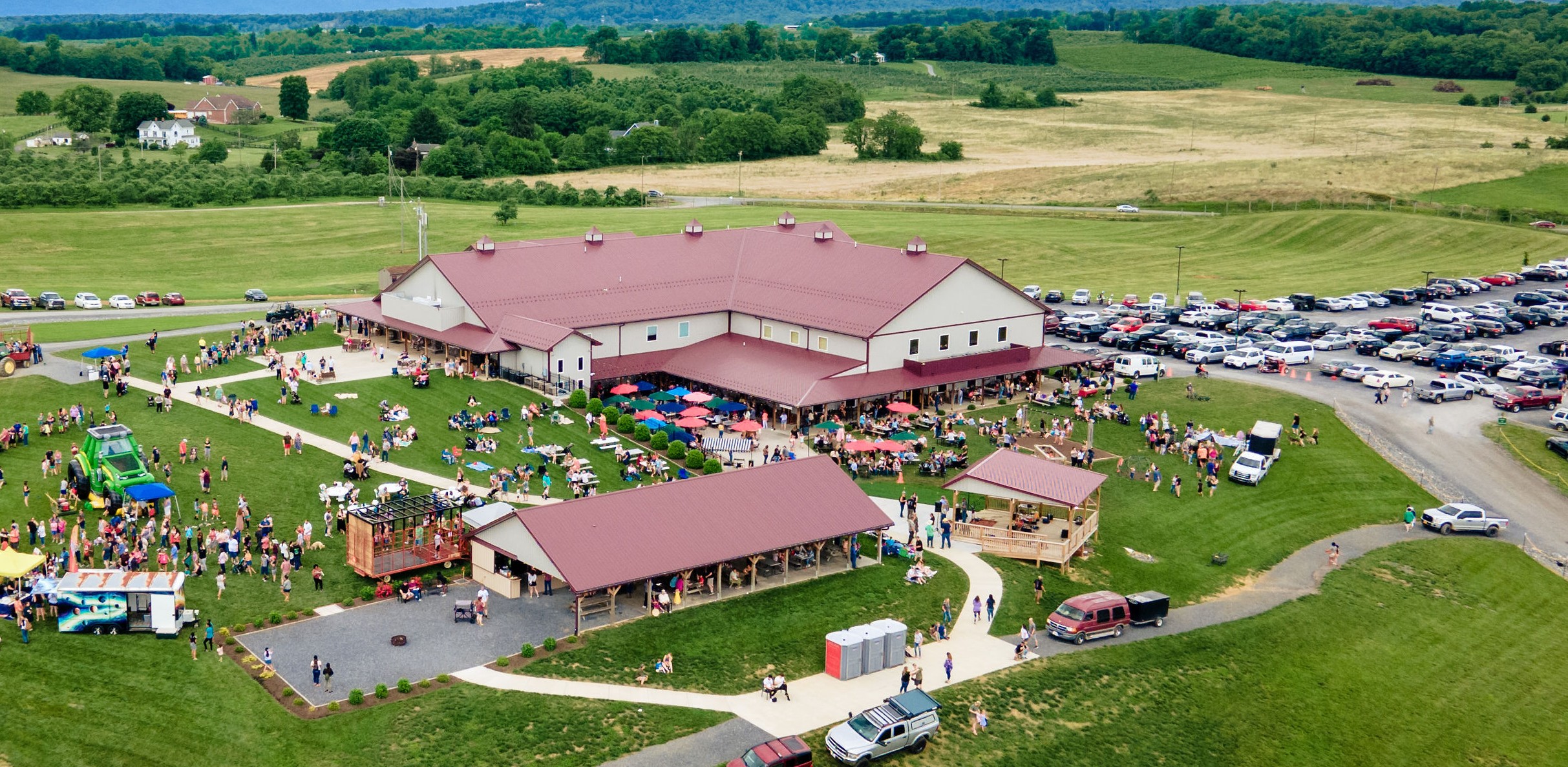 Large outdoor event with tents, people, and a building in a green field, surrounded by trees and open land.