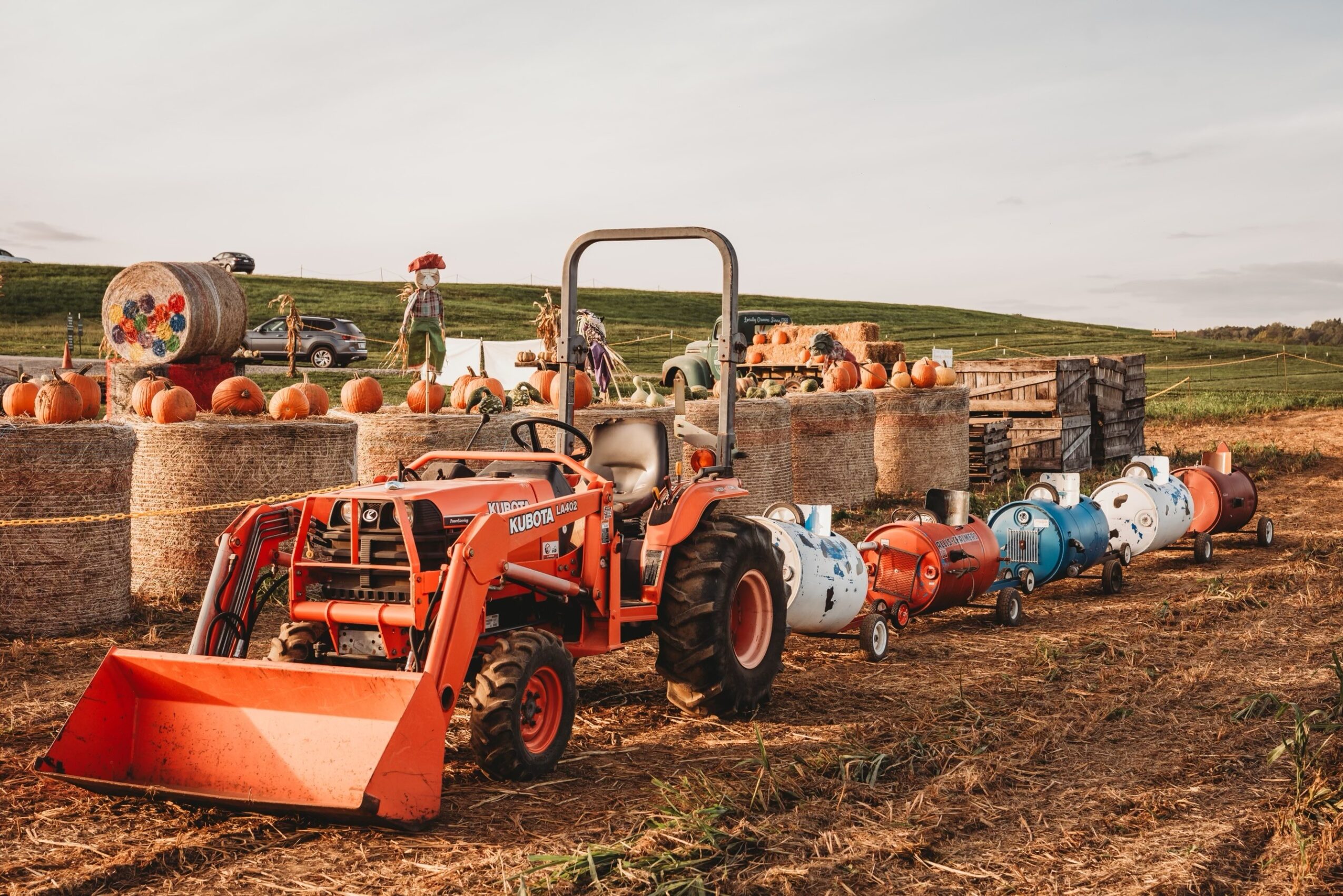 Farm scene with hay bales, pumpkins, and a red tractor pulling a trailer loaded with barrels on a field.