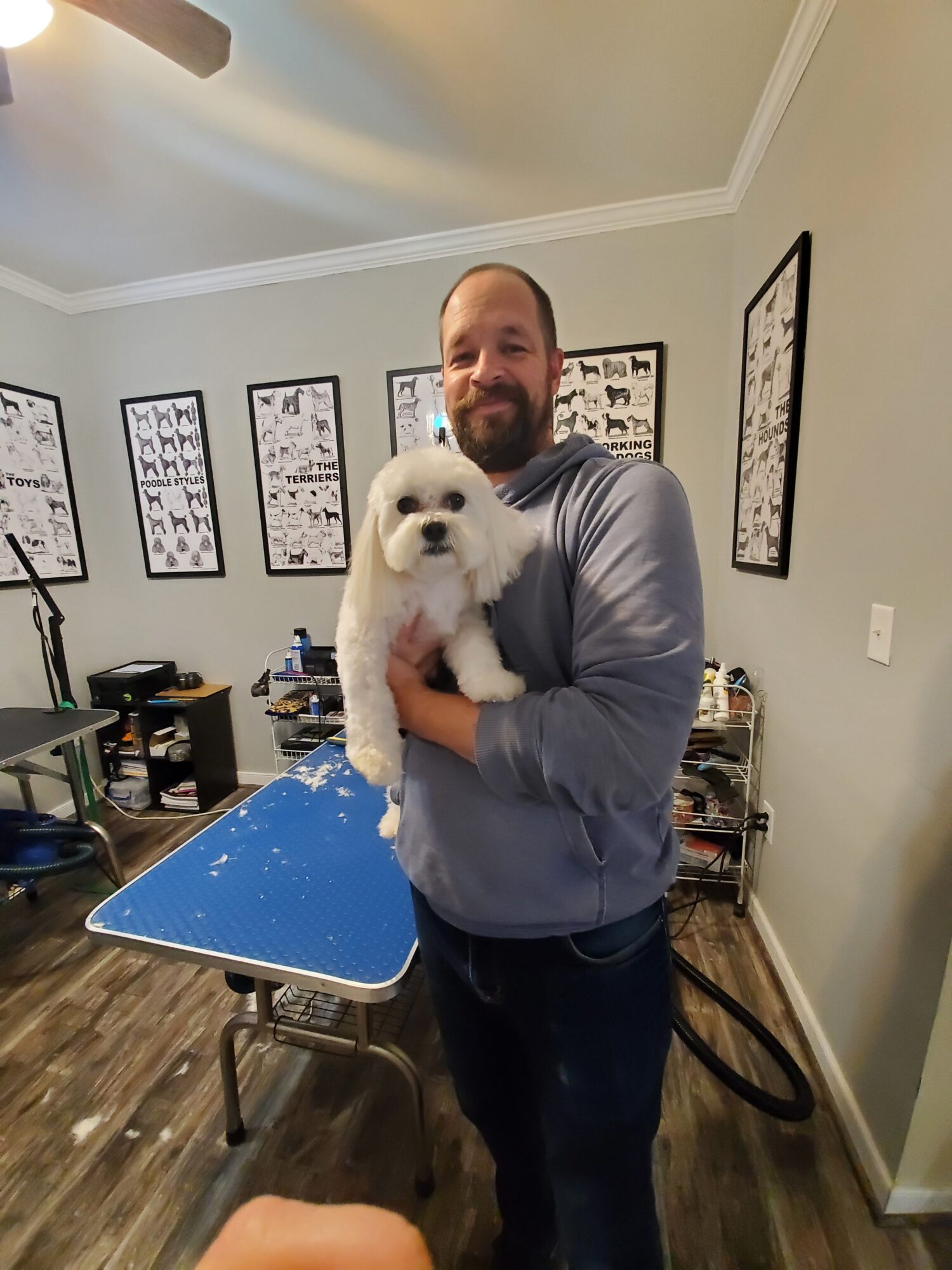 Man holding a white puppy indoors, with framed pictures on the wall behind him.