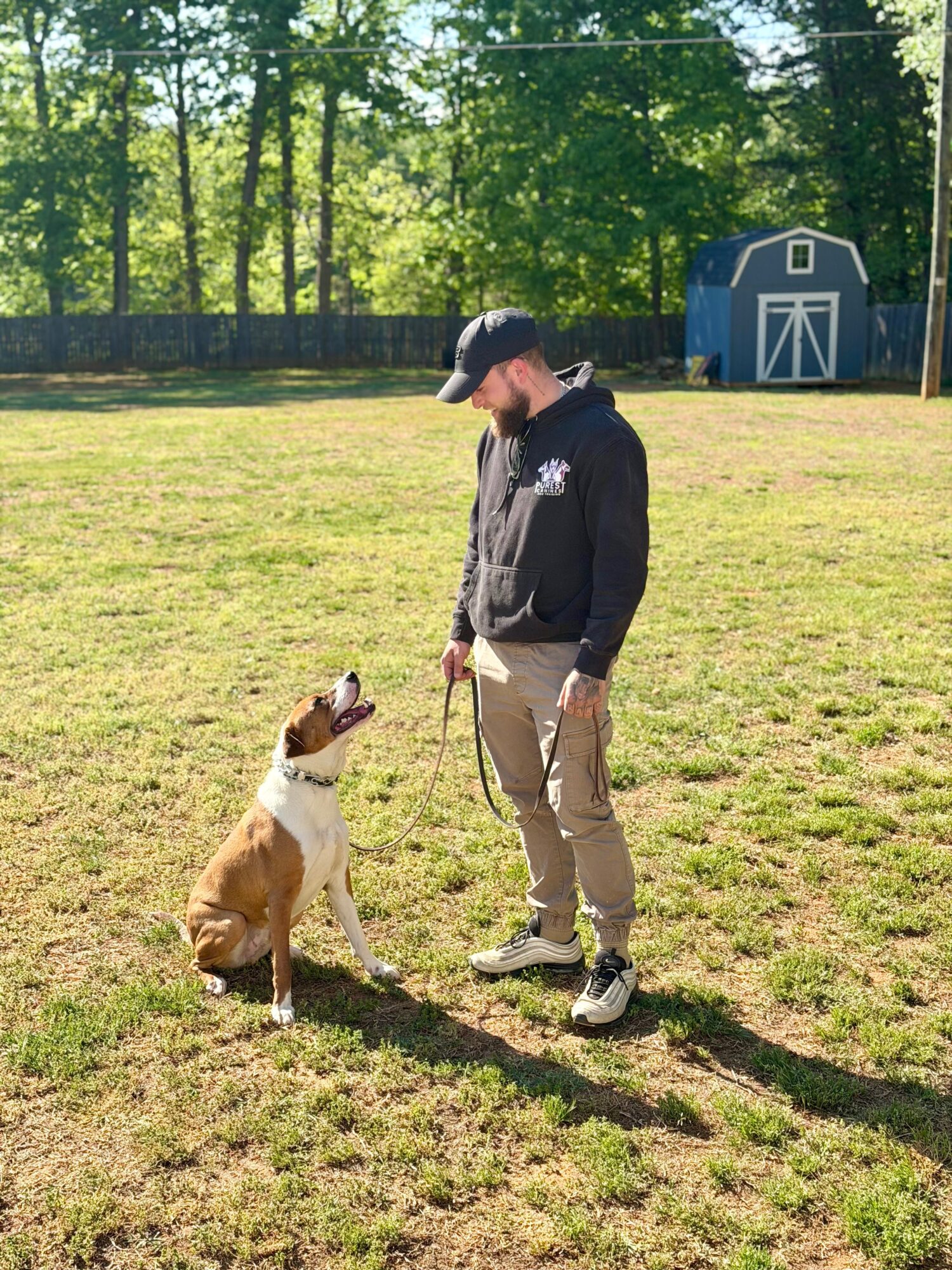 Person in black hoodie and beige pants with a dog sitting on grass, trees and a blue shed in background.
