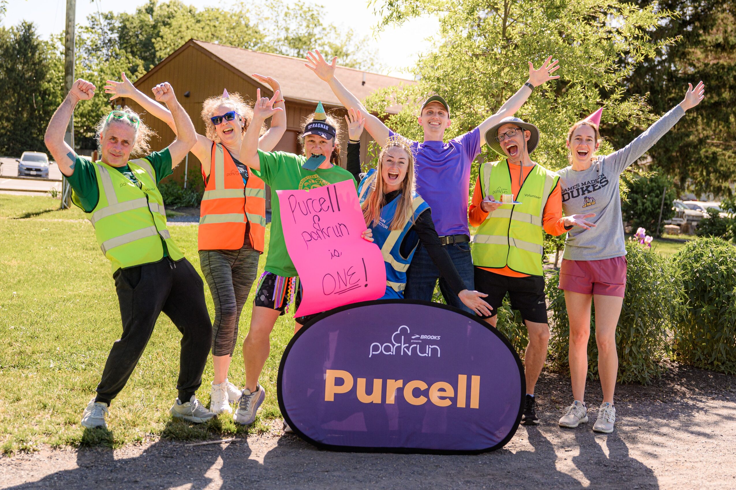 Group of children outdoors holding a pink sign and standing behind a purple Purcell-branded bag, smiling and raising hands.