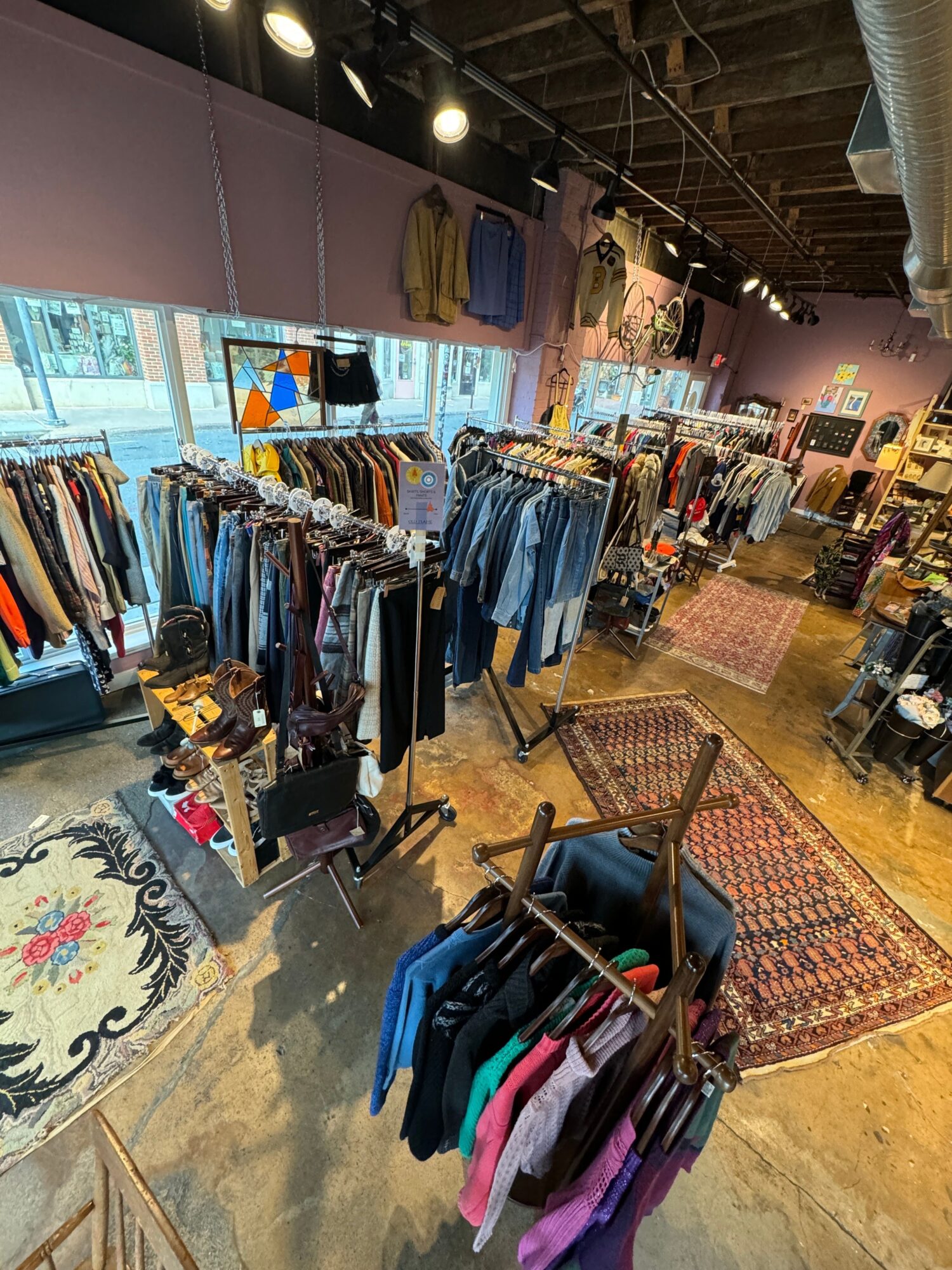 Clothing store interior with racks of jeans, shirts, and accessories, wooden floors, and a patterned rug near the entrance.