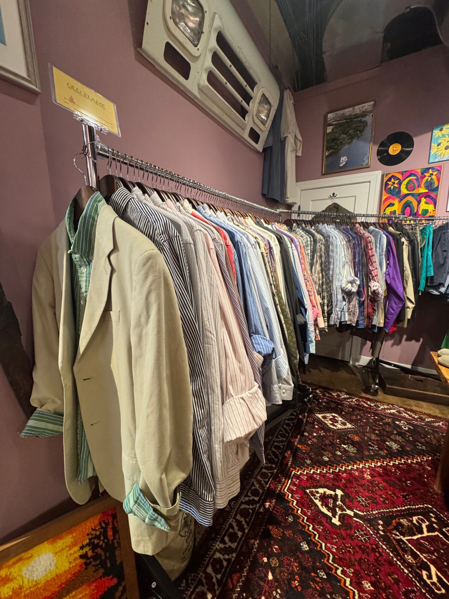 Clothing and fabric items arranged on a table with a patterned rug nearby, pink wall, and artwork in the background.