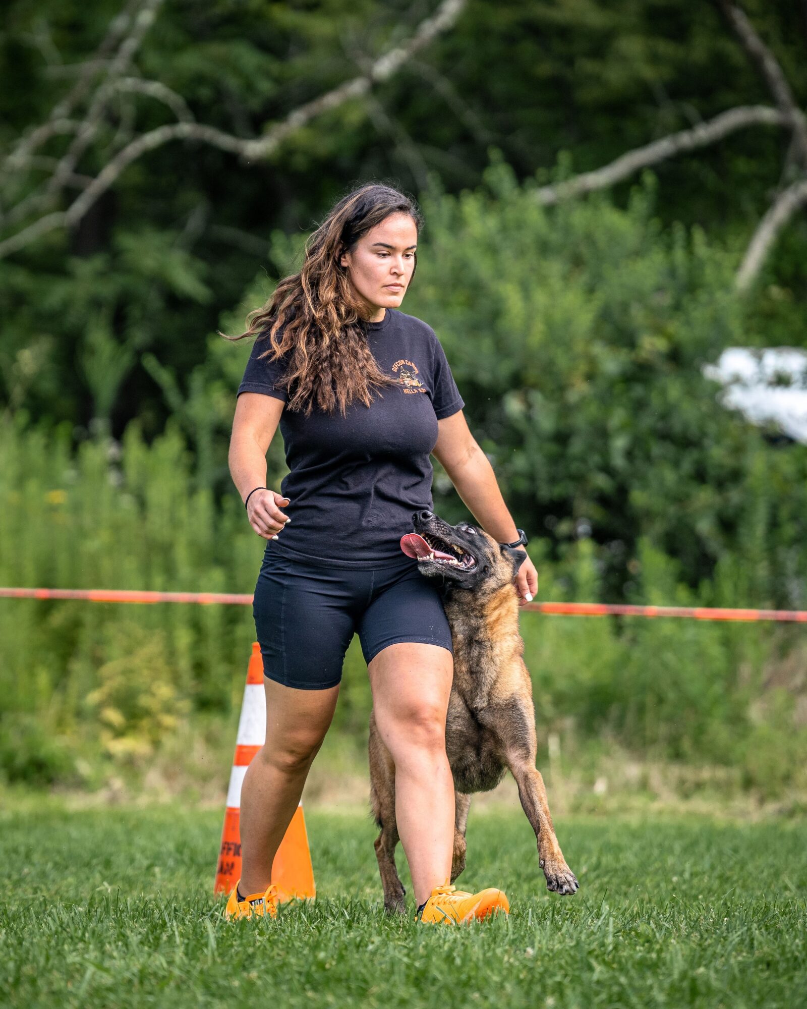 Young woman standing outdoors with a Belgian Malinois dog, green trees and grass in background, orange traffic cone nearby.