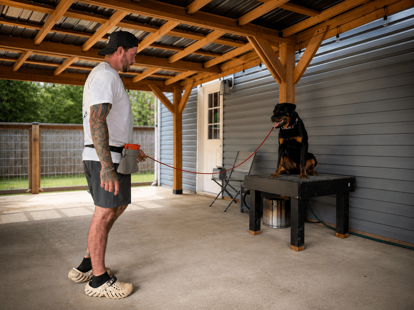Man standing on concrete under a wooden roof, holding a leash attached to a dog on a table outside a house.