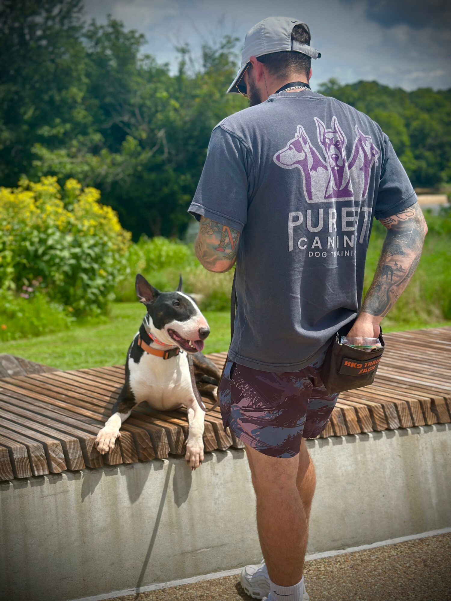 Man with tattoos and a cap standing outdoors next to a dog on a leash, with greenery in the background.