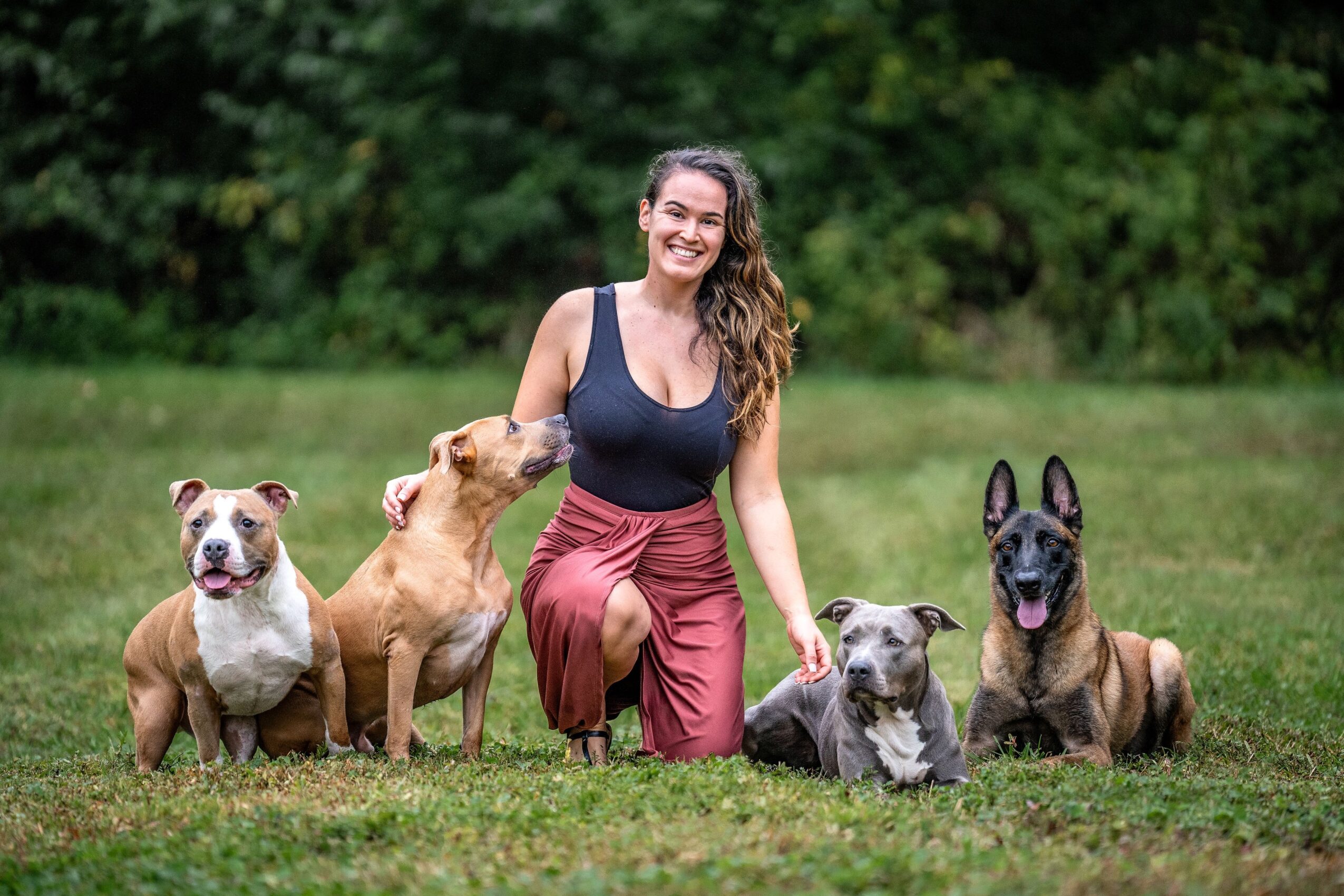Woman kneeling on grass with five dogs outdoors, trees in background, smiling, wearing black top and red pants.