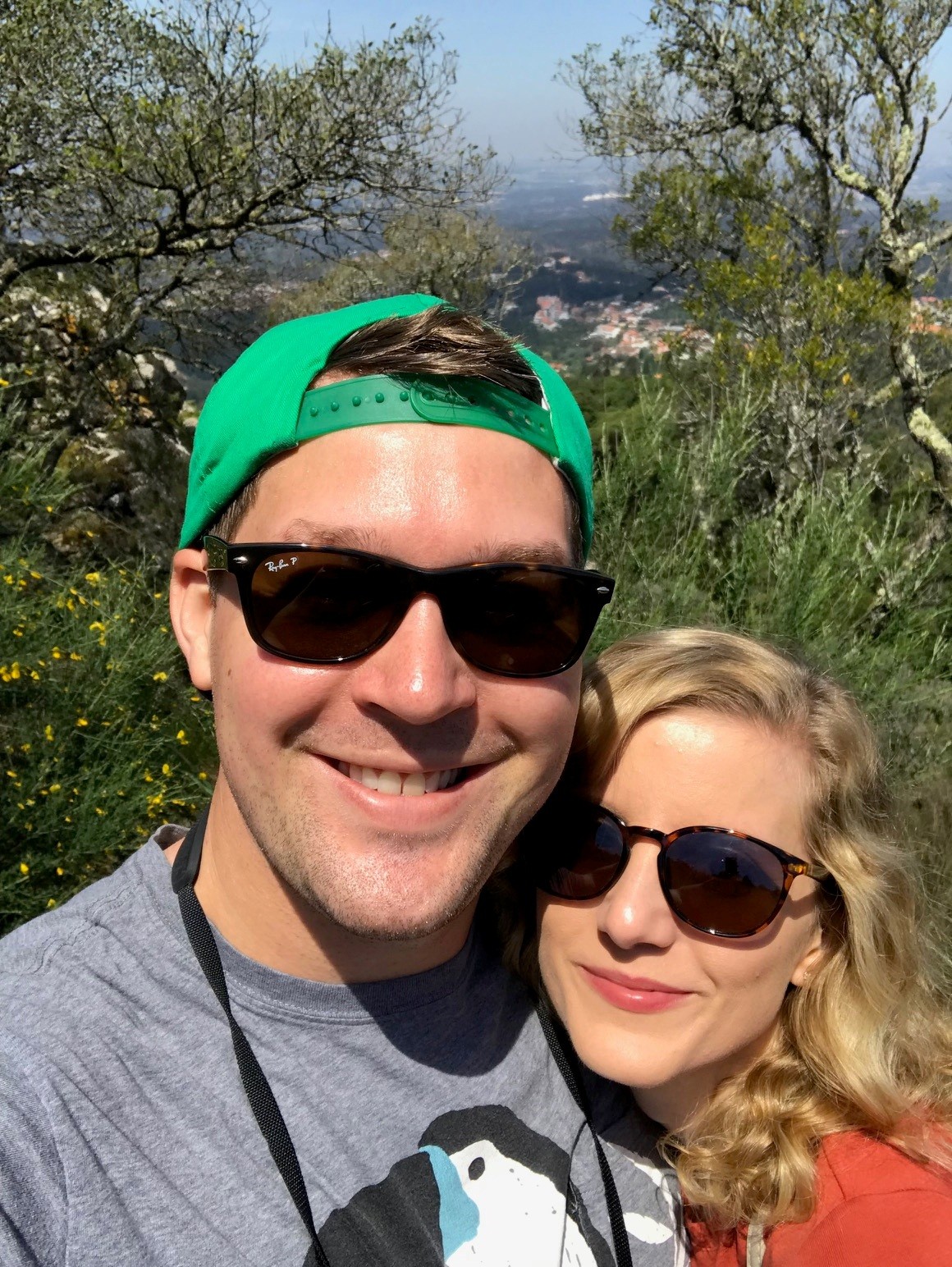 A smiling man and woman wearing sunglasses outdoors with trees and a view in the background.