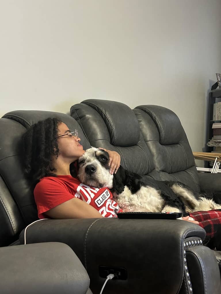 Child with glasses and curly hair sitting on a black leather couch, holding a black and white dog, in a living room.