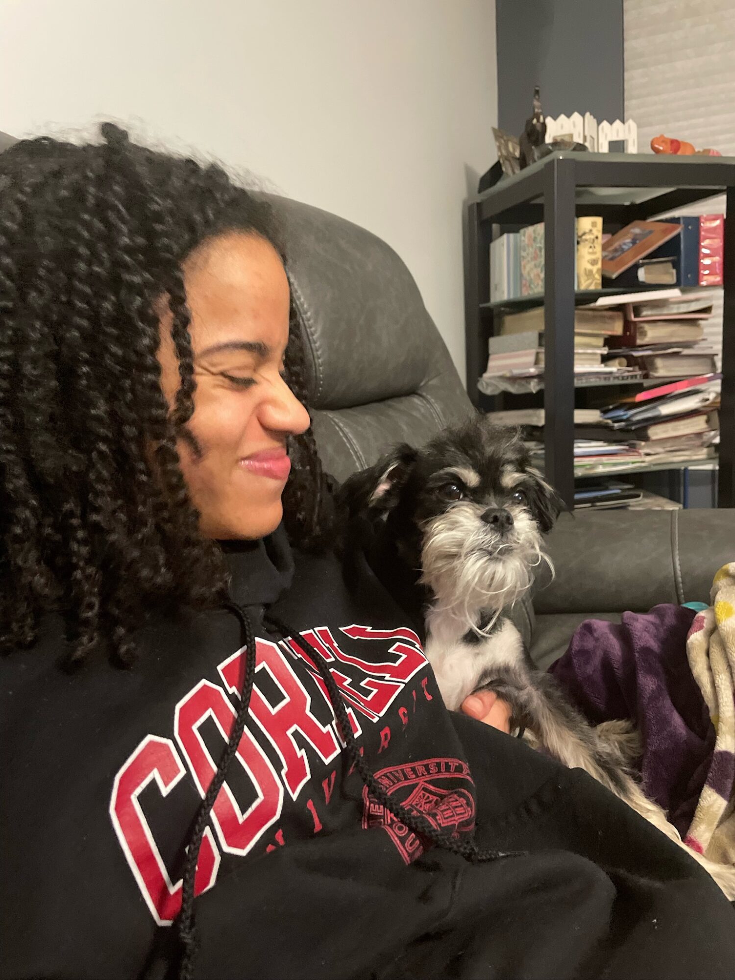 Person with curly hair smiling while holding a small black and white dog on their lap, sitting on a couch.