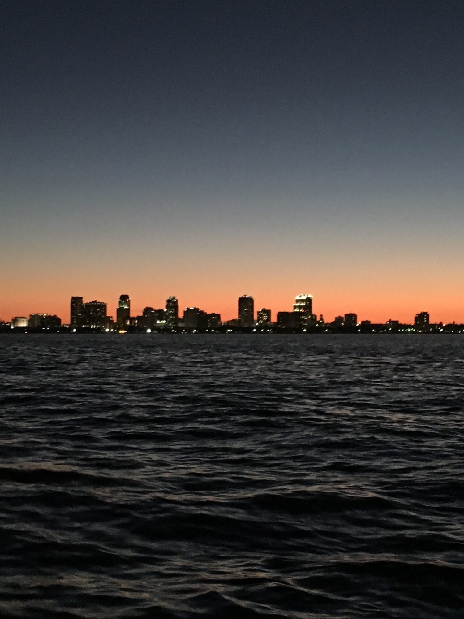 City skyline silhouette at sunset with water in foreground, dark sky above, and orange glow near horizon.