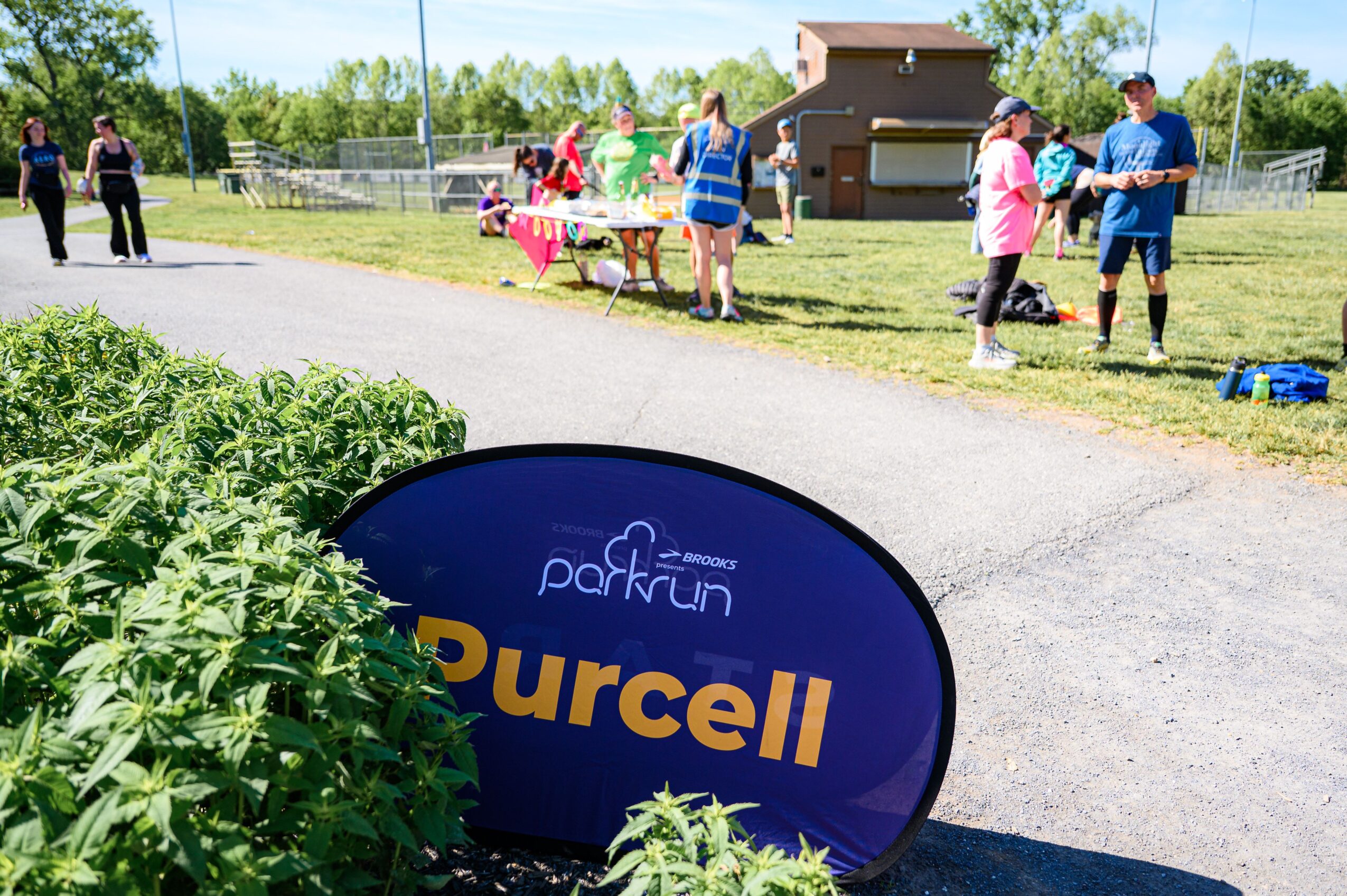 People gather outdoors near a building and tables, with a sign for Purcell park run in the foreground.