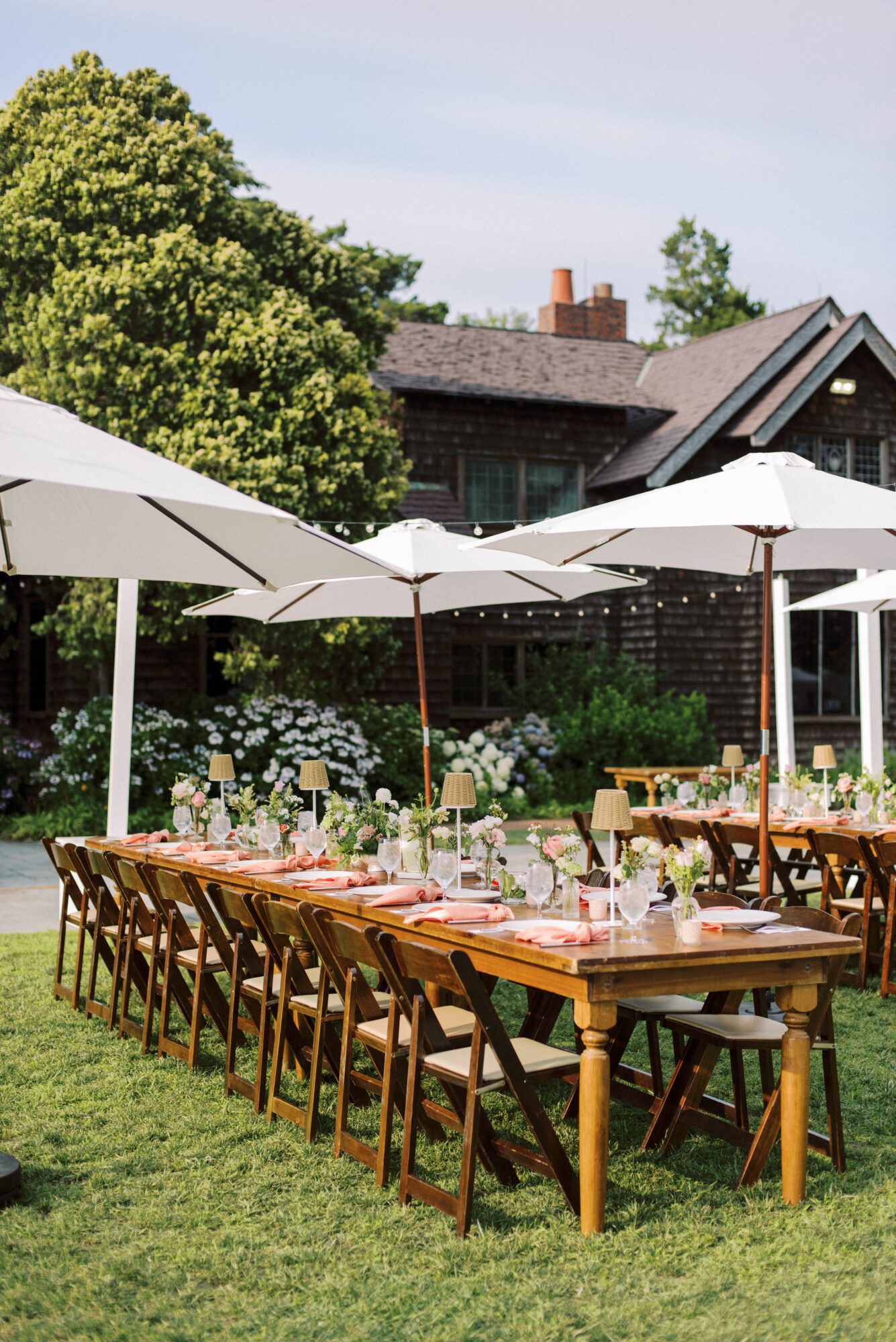 Long outdoor table with chairs and white umbrellas in a garden setting, with trees and houses in the background.