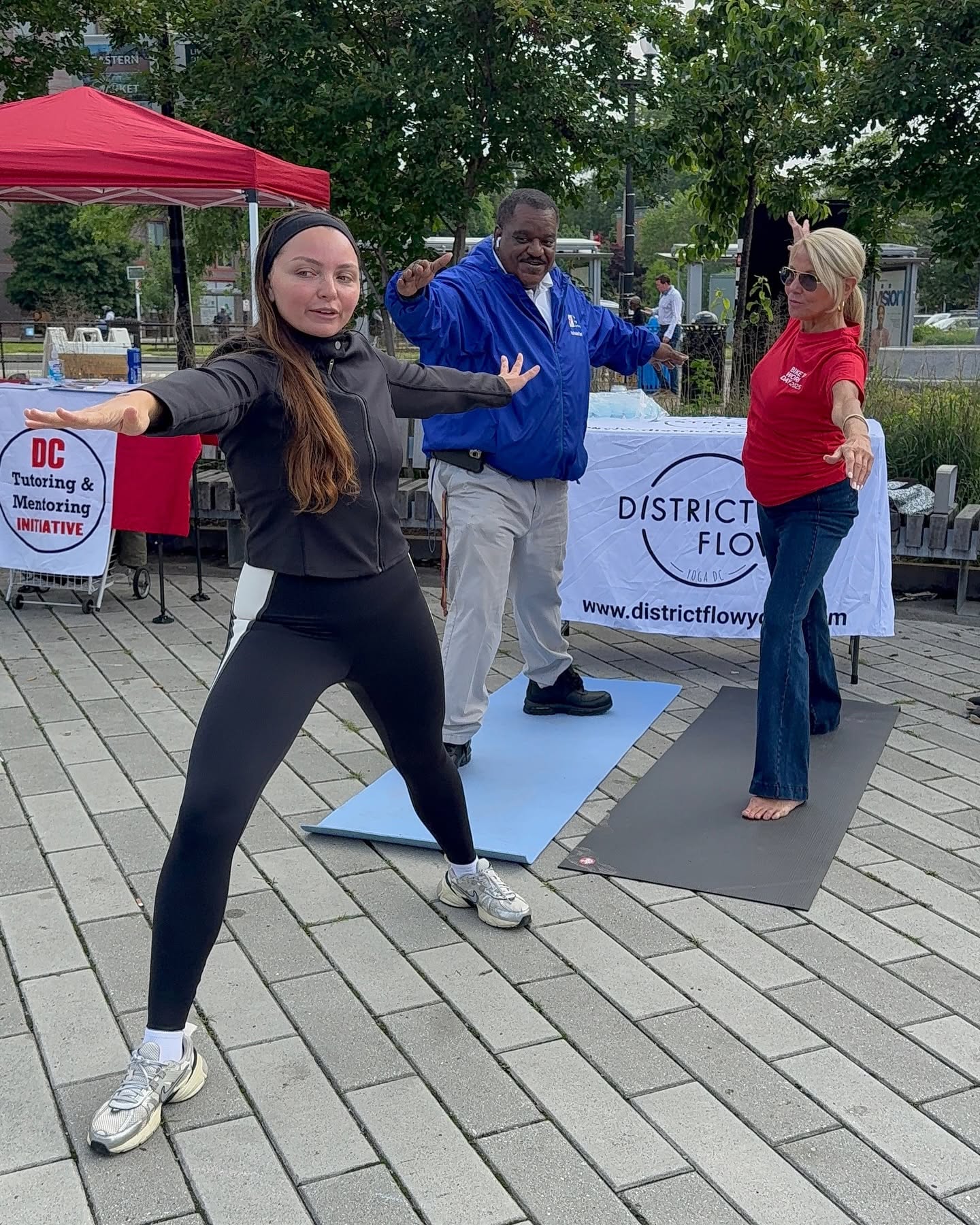 Three people participating in outdoor yoga or stretching session on a paved area with trees and tents in background.