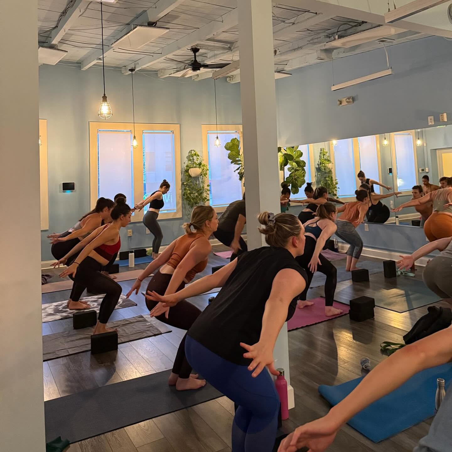 Group of people participating in a yoga class in a bright studio with large windows and plants.