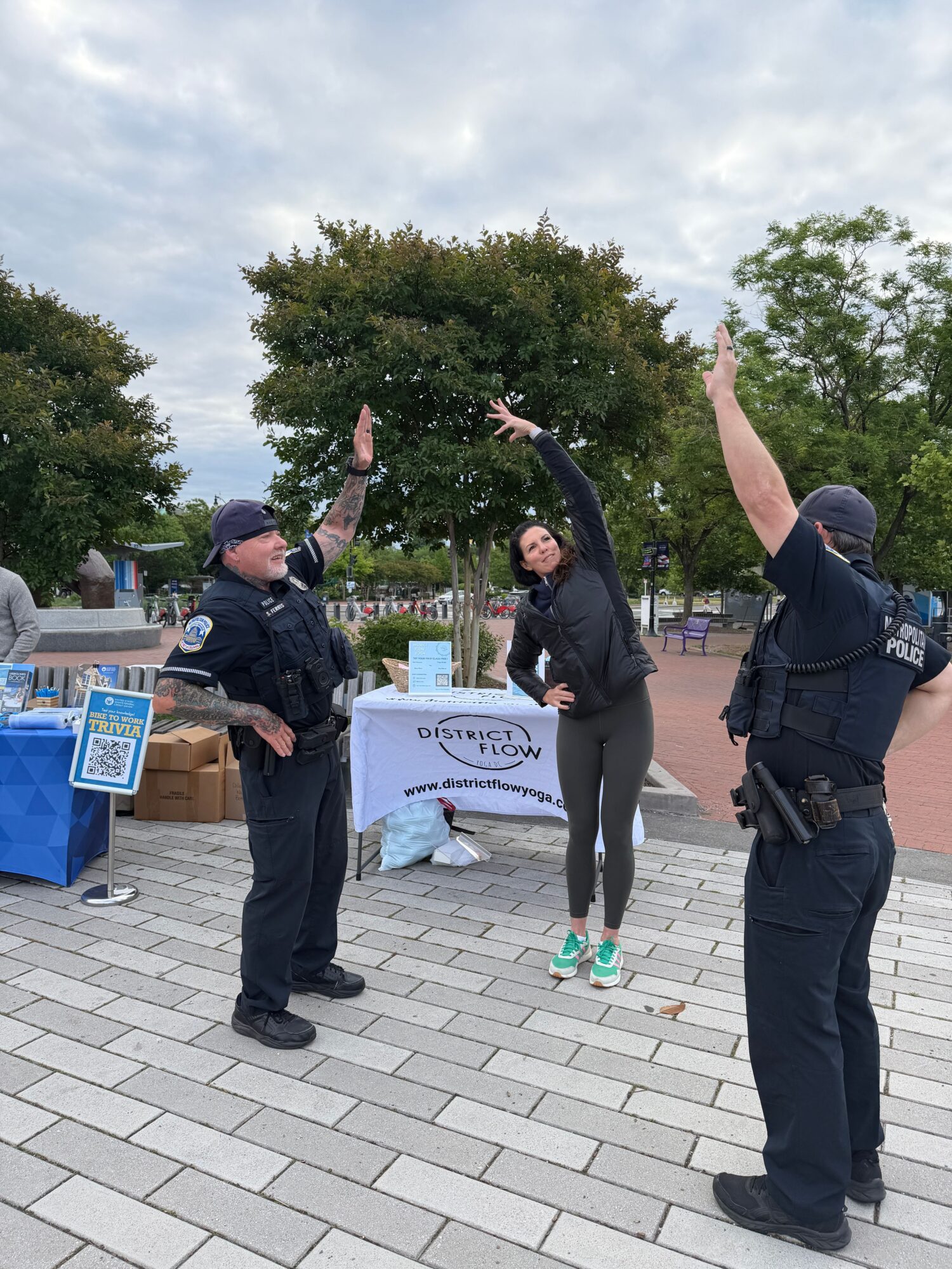 Two police officers and a person with a skateboard near a table with signs and supplies outdoors, trees in background.