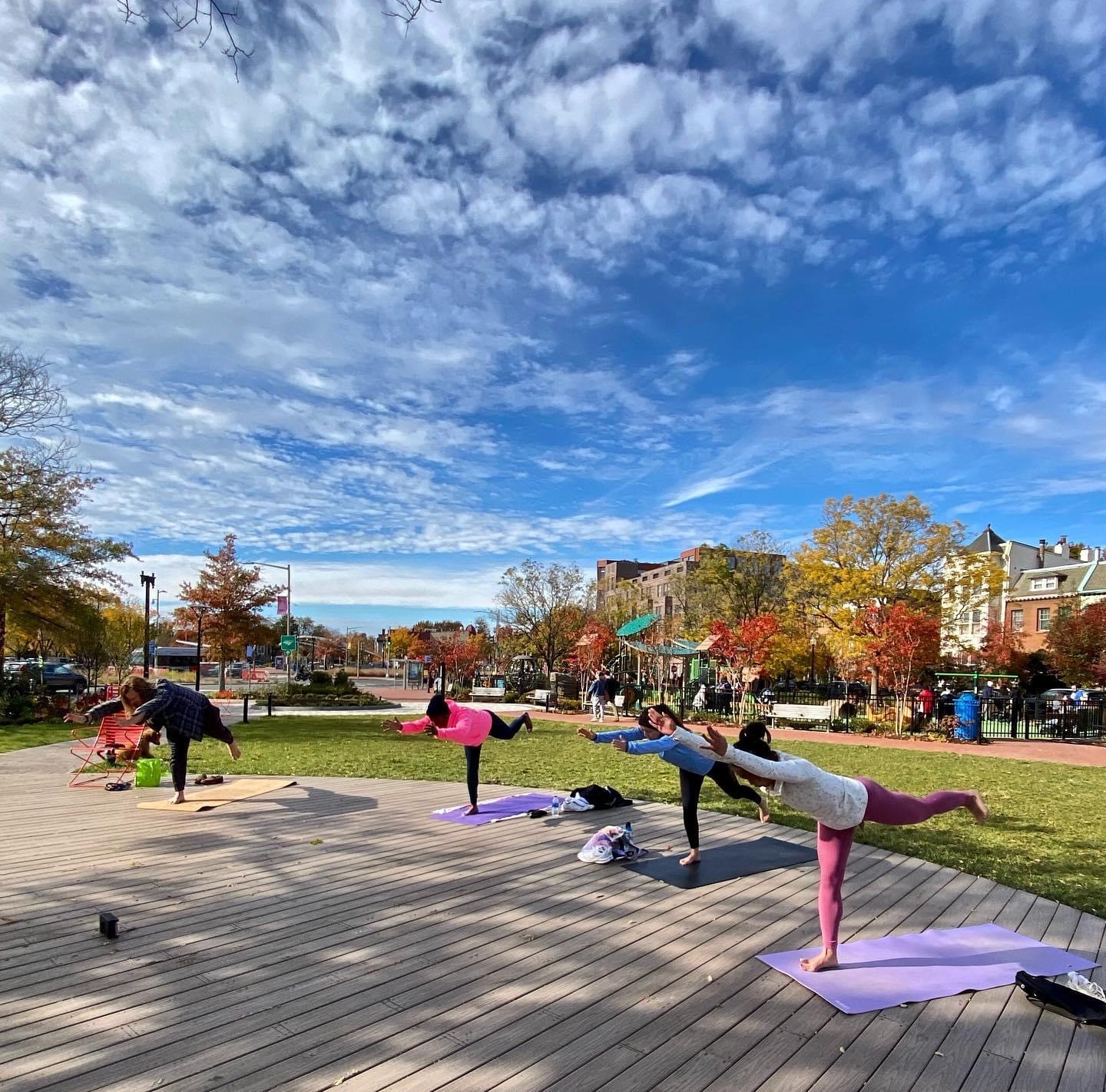 Group of people practicing yoga outdoors on mats in a park with trees and buildings, under a partly cloudy sky.