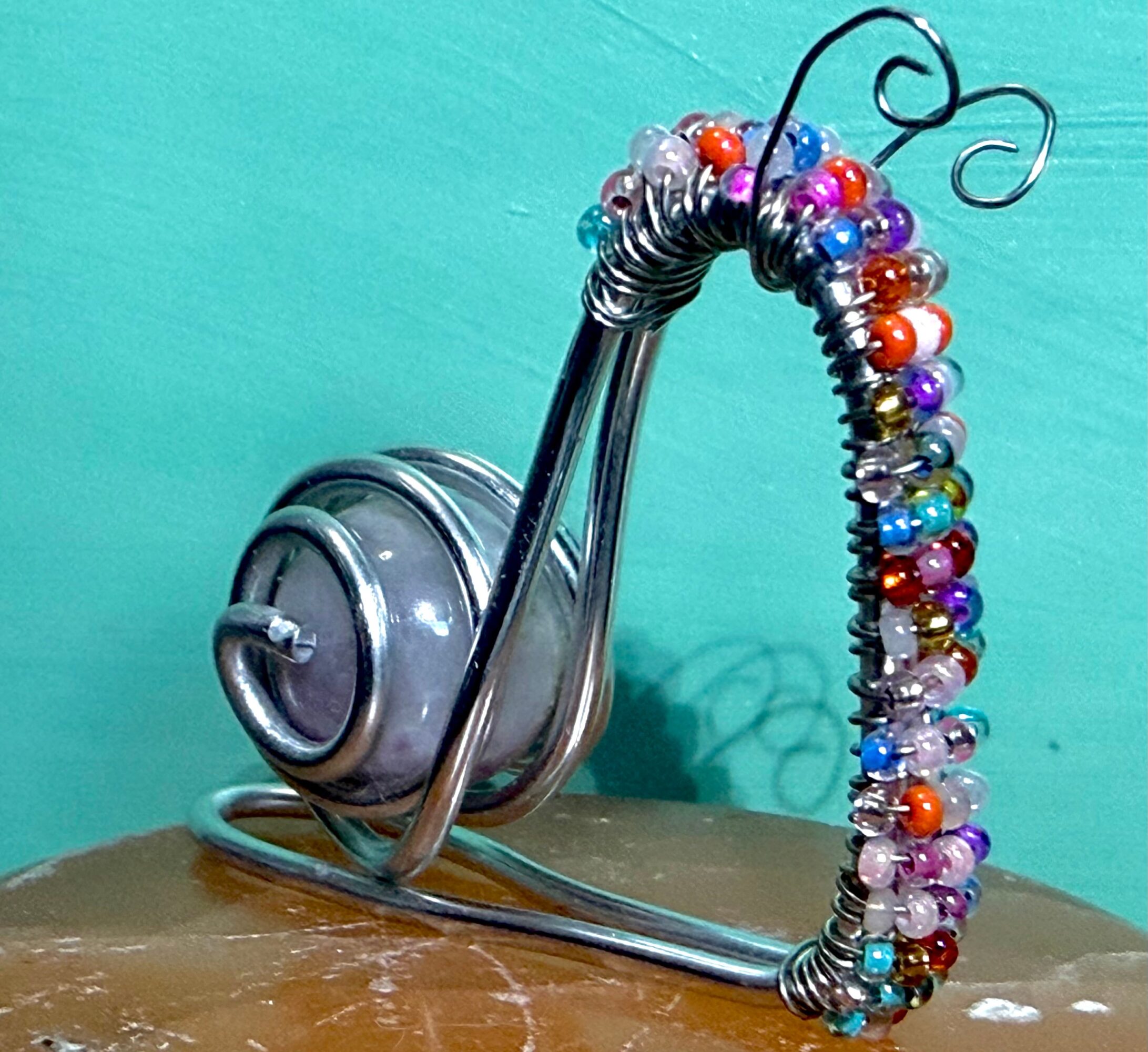 Wire snail sculpture with colorful beads on a wooden surface against a teal background.
