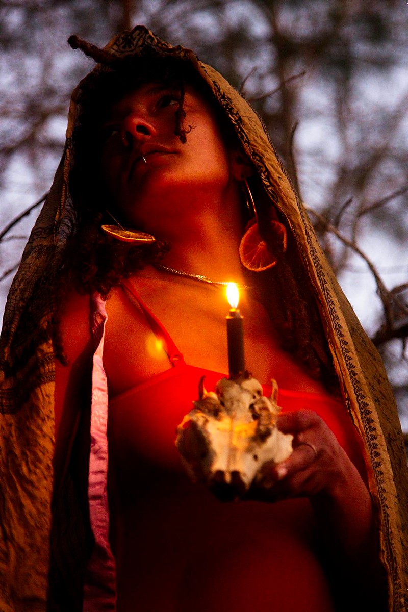 Woman holding a lit candle and a small animal, with a dark background and trees, wearing a head covering.
