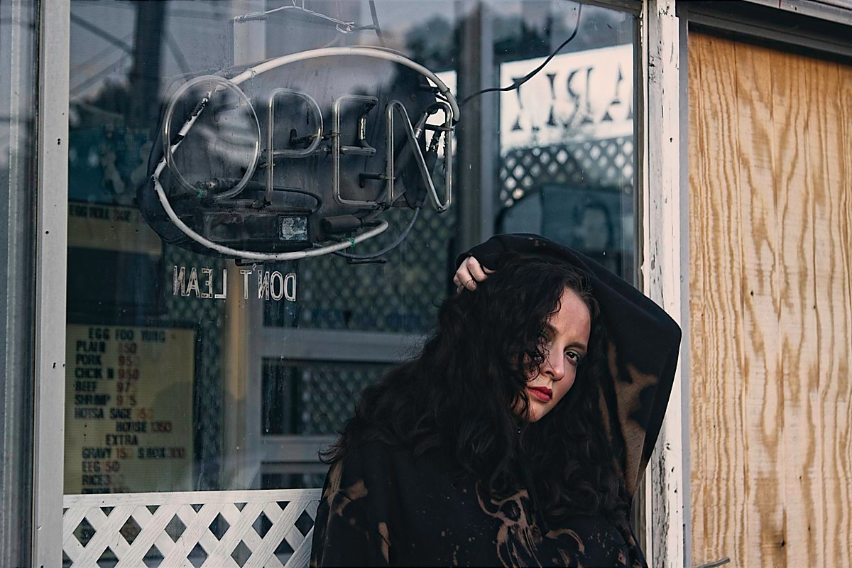 Woman with dark curly hair standing outside a shop with an open sign, holding her head with one hand.