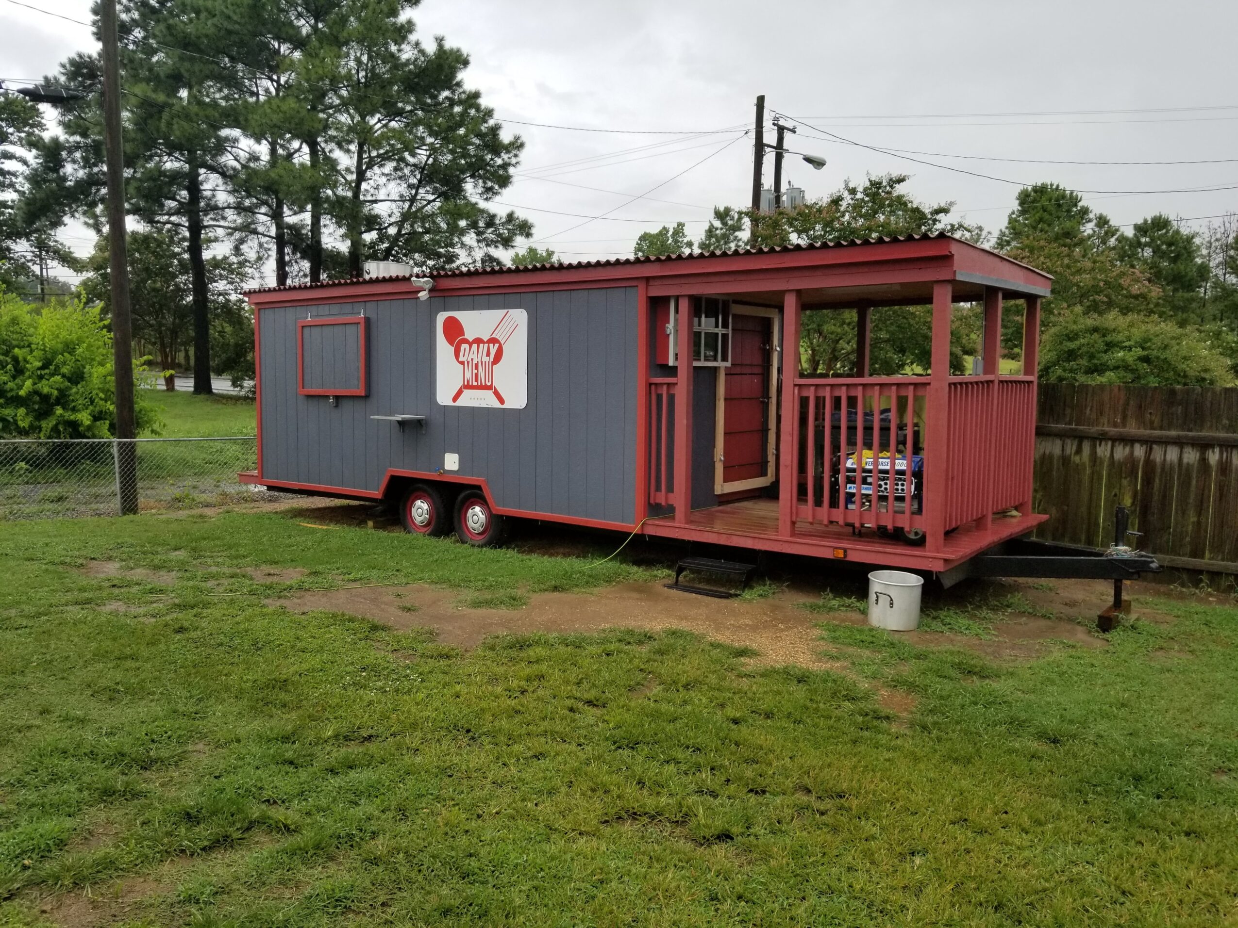 A small, gray and red trailer with a porch, set on grass with trees and a fence in the background.
