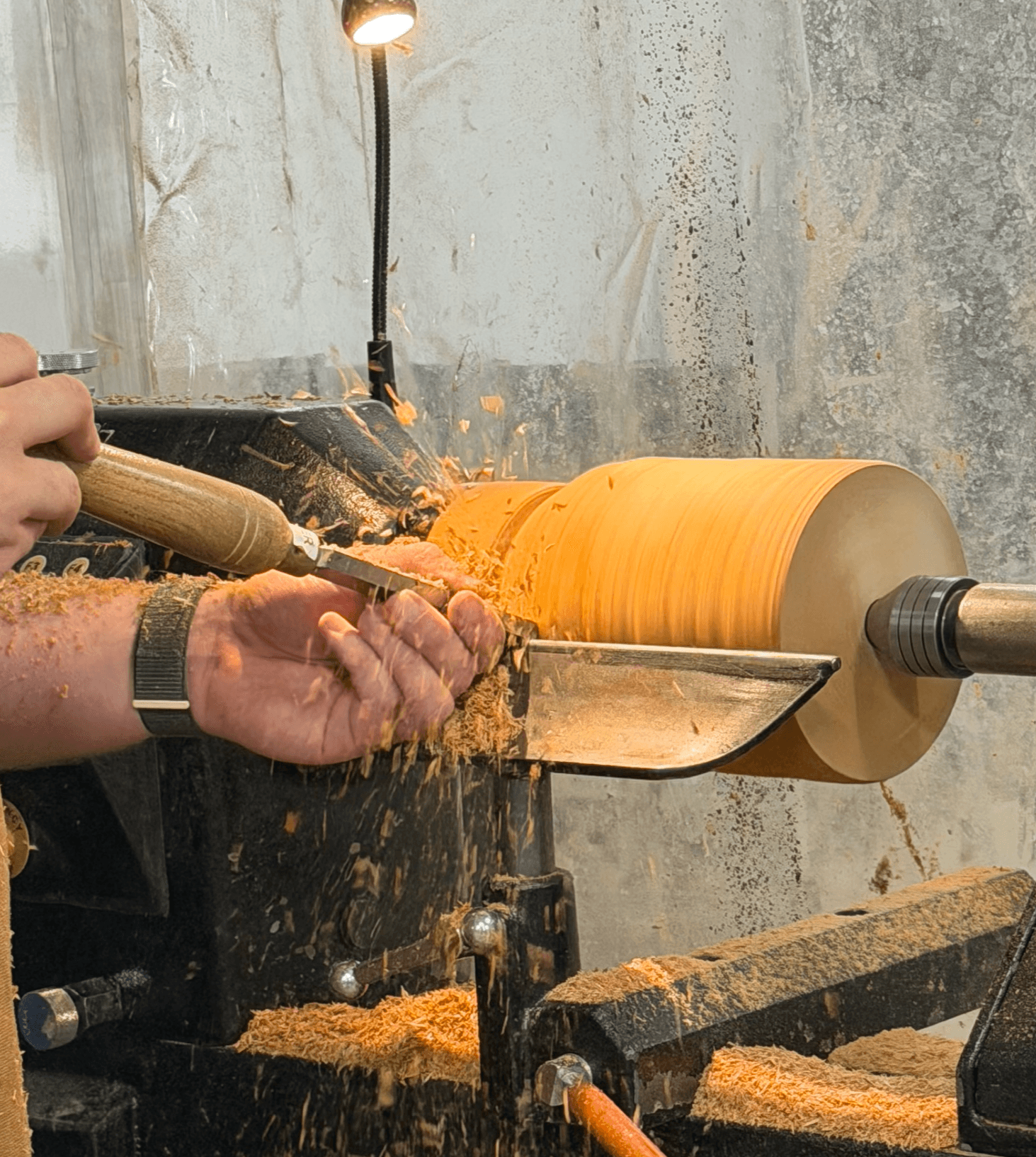 Person shaping a wooden bowl on a lathe with wood shavings around, illuminated by a small lamp, in a workshop.