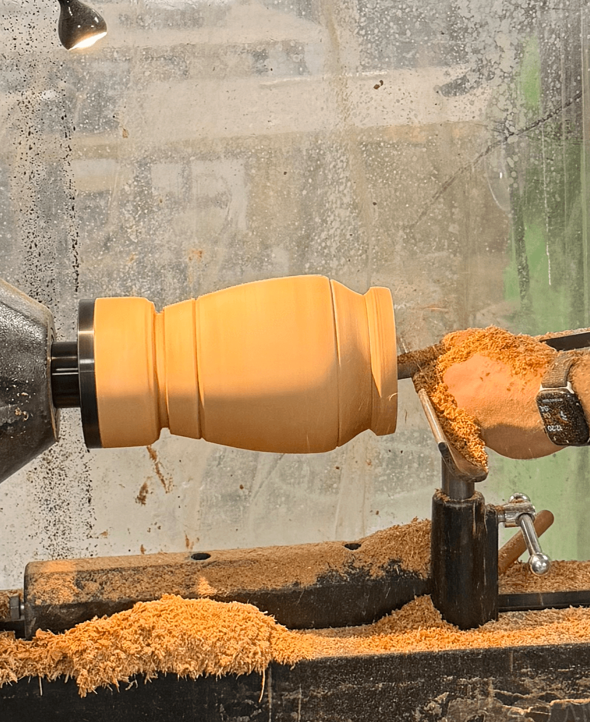 Woodturning lathe with a partially shaped wooden piece, shavings on the lathe bed, and a workshop background.